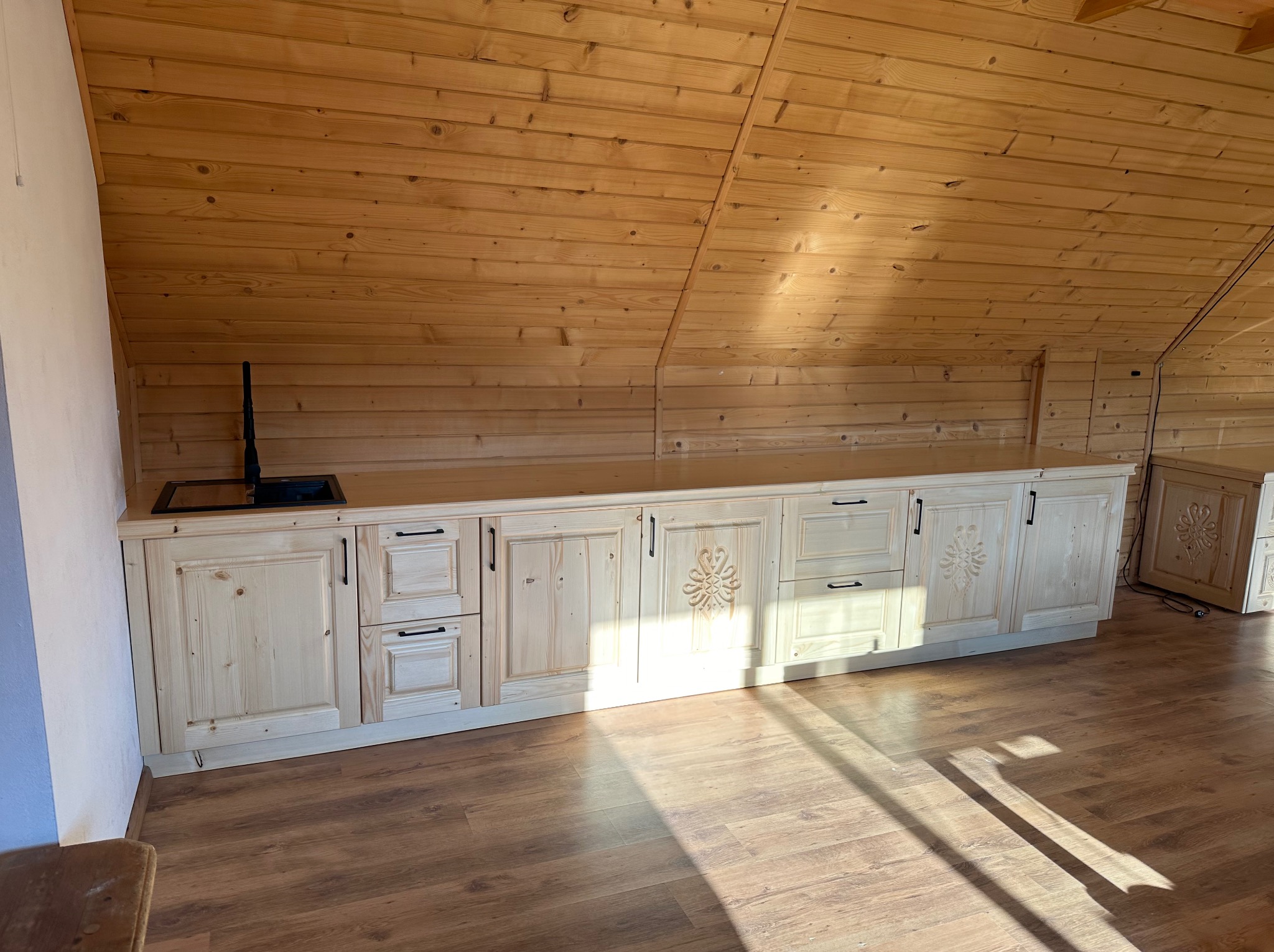 Jasne drewniane szafki kuchenne z czarnymi uchwytami i dekoracyjnymi rzeźbieniami w kształcie kwiatów, umieszczone na poddaszu z drewnianą boazerią. Widoczny czarny zlewozmywak i drewniana podłoga.