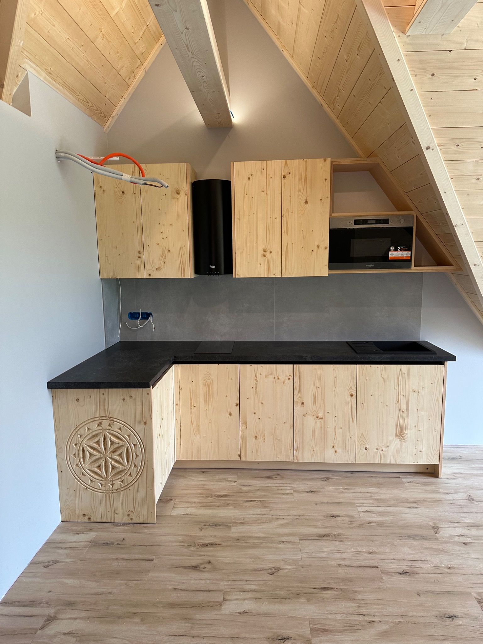 Drewniana kuchnia na poddaszu, front szafki z unikalnym wzorem rozety, czarny blat, sprzęt AGD. Widoczne drewniane wykończenie poddasza.