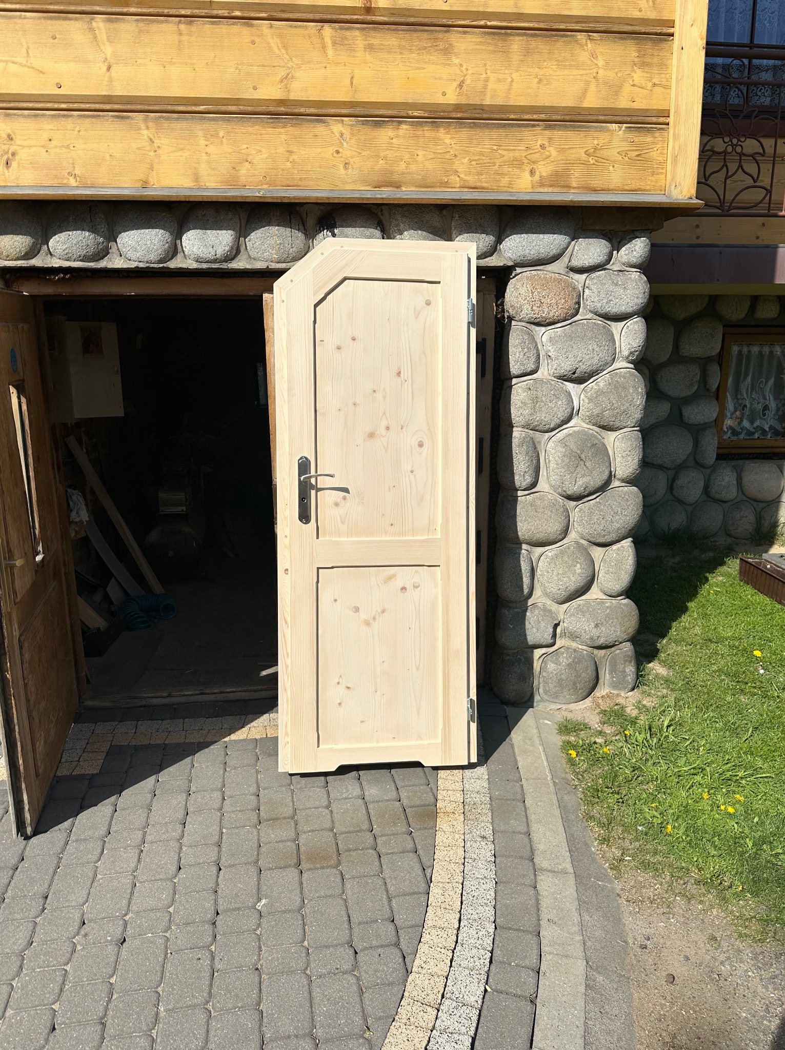 Otwarta, surowa drewniana dwuskrzydłowa brama z czarną klamką, osadzona w kamiennej ścianie budynku z drewnianym wykończeniem, na tle brukowanej ścieżki i trawy.