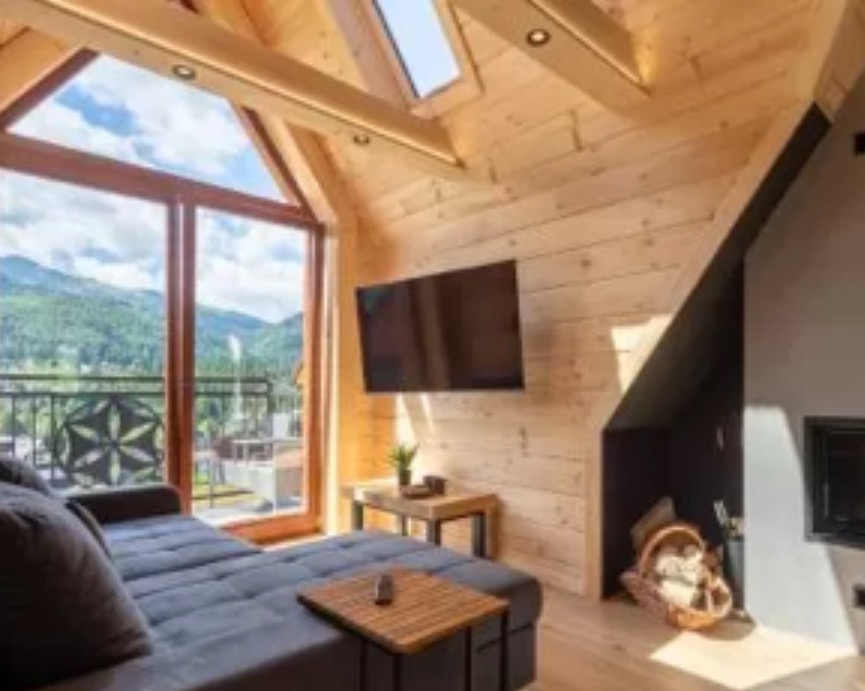 Drewniane wnętrze w stylu góralskim z dużym oknem balkonowym i widokiem na góry. Drewniane ściany, sufit ze świetlikami i nowoczesny telewizor.