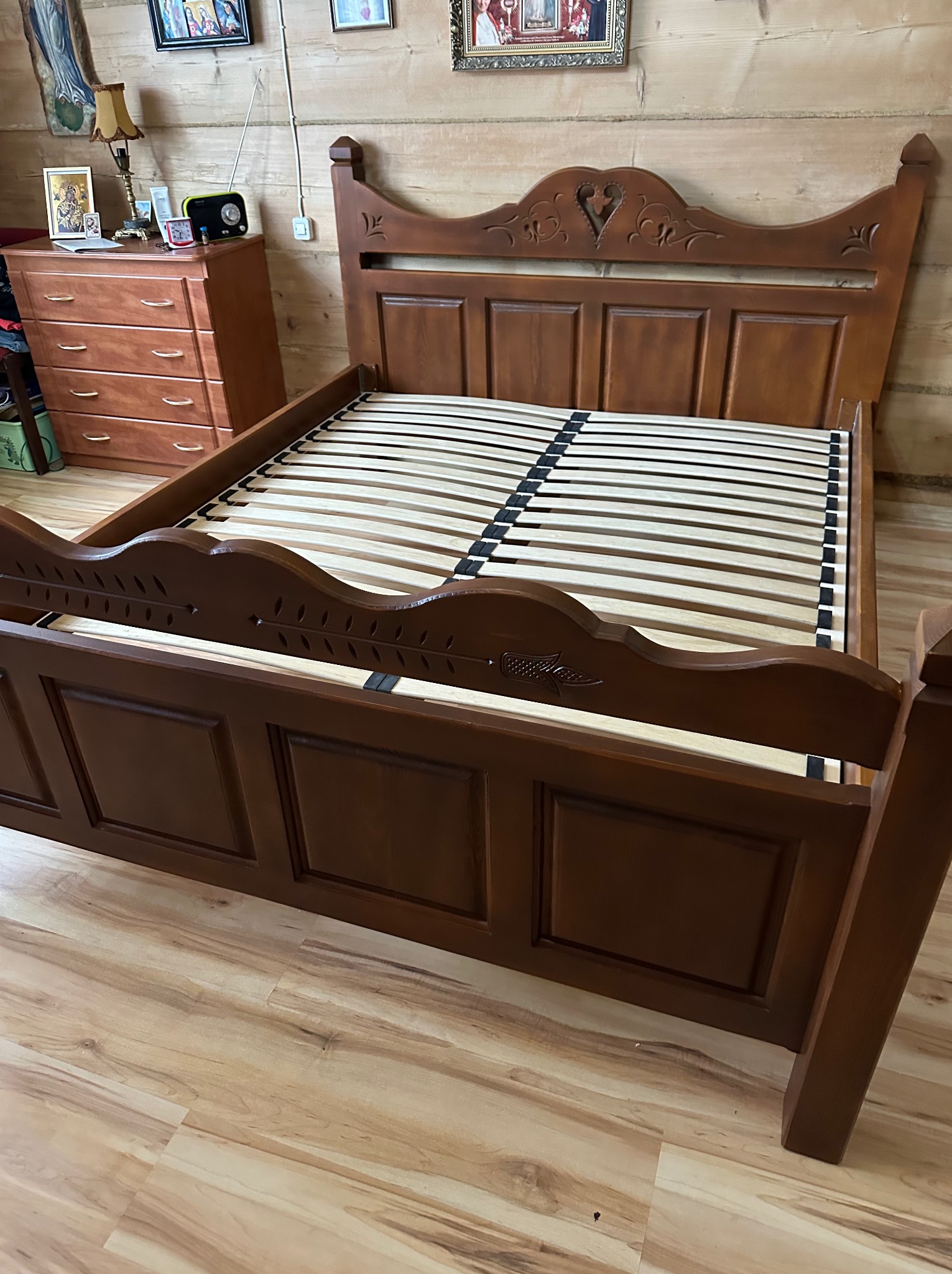 Drewniane łóżko z dekoracyjnym zagłówkiem w rustykalnym wnętrzu. Widoczna drewniana komoda w tle. Łóżko z listwowym stelażem, brązowe wykończenie drewna.