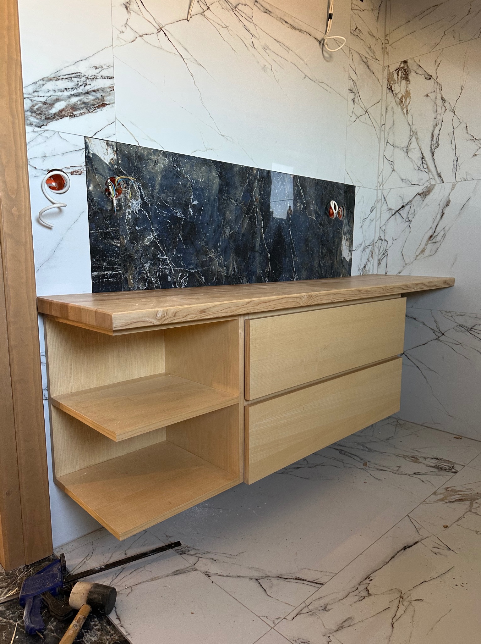 Drewniana szafka łazienkowa z blatem z jasnego drewna, z otwartymi półkami i szufladami, na tle marmurowych płytek. Widoczne narzędzia stolarskie na podłodze.