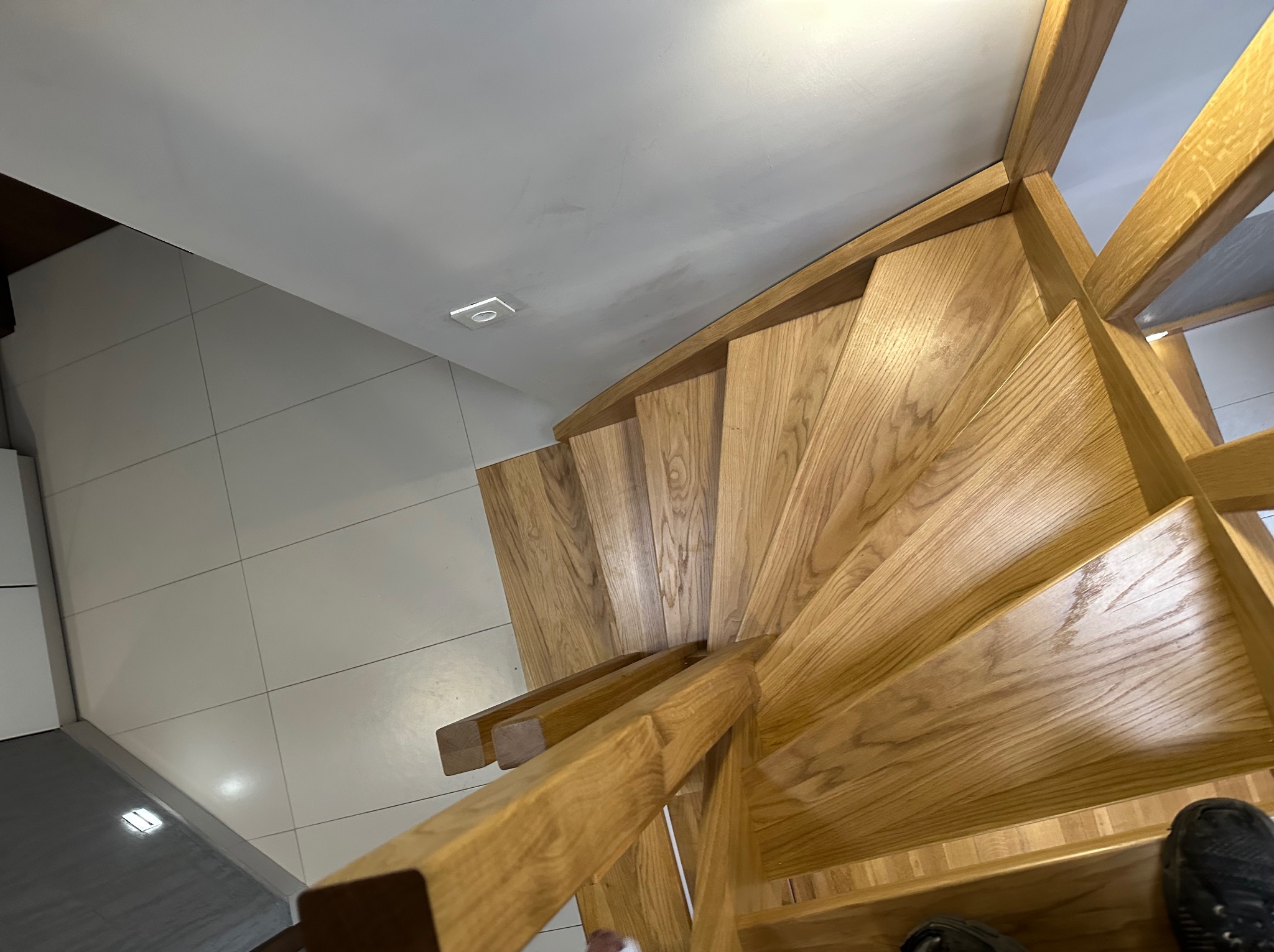 Drewniane schody spiralne z jasnego drewna dębowego, widoczne z góry, z fragmentem białej podłogi i sufitu. U dołu kadru fragment buta.