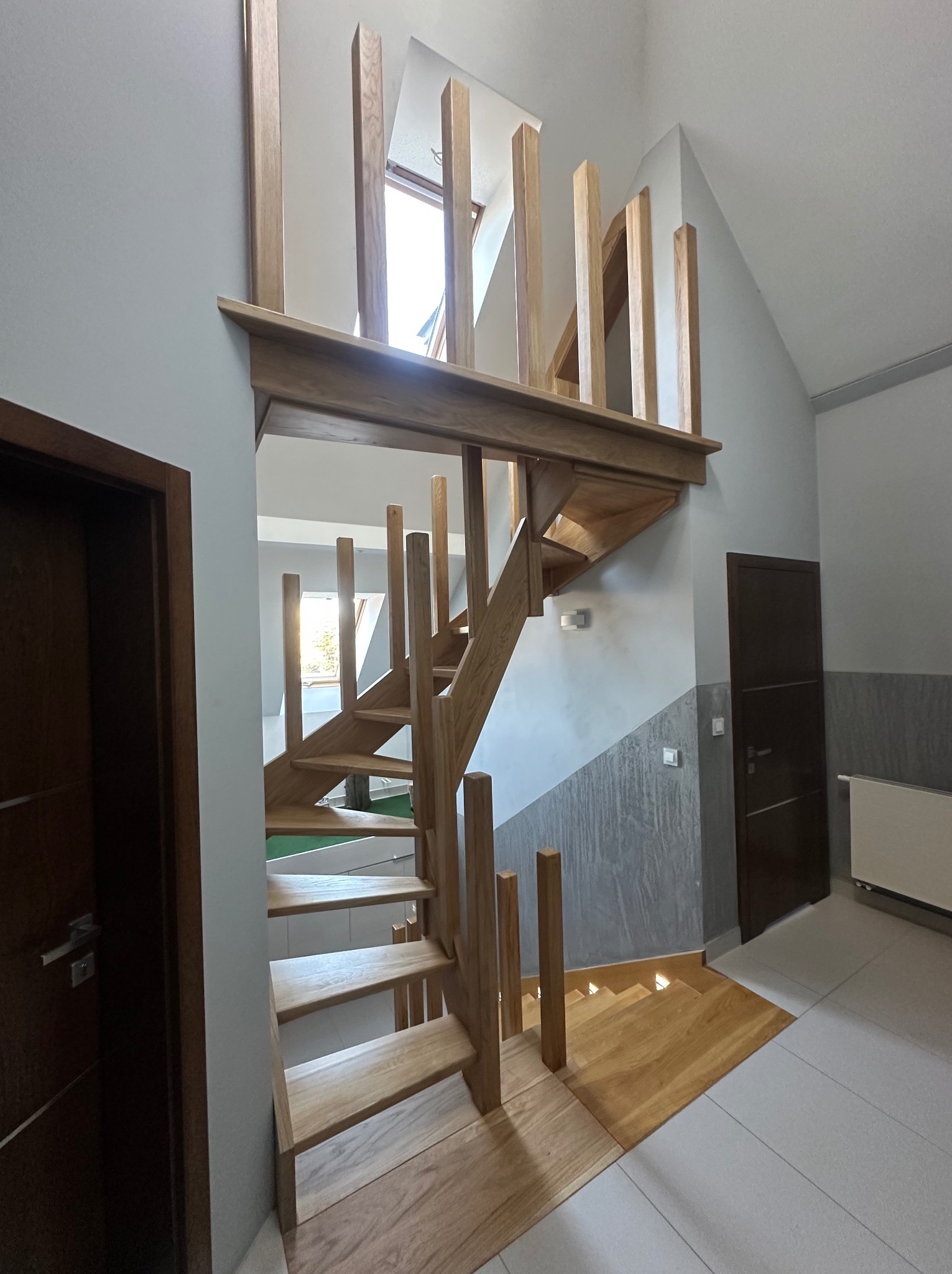 Drewniane schody z jasnego drewna, z pionowymi słupkami zamiast balustrady, prowadzące na antresolę z oknem dachowym. Minimalistyczny design, nowoczesne wykończenie wnętrza.