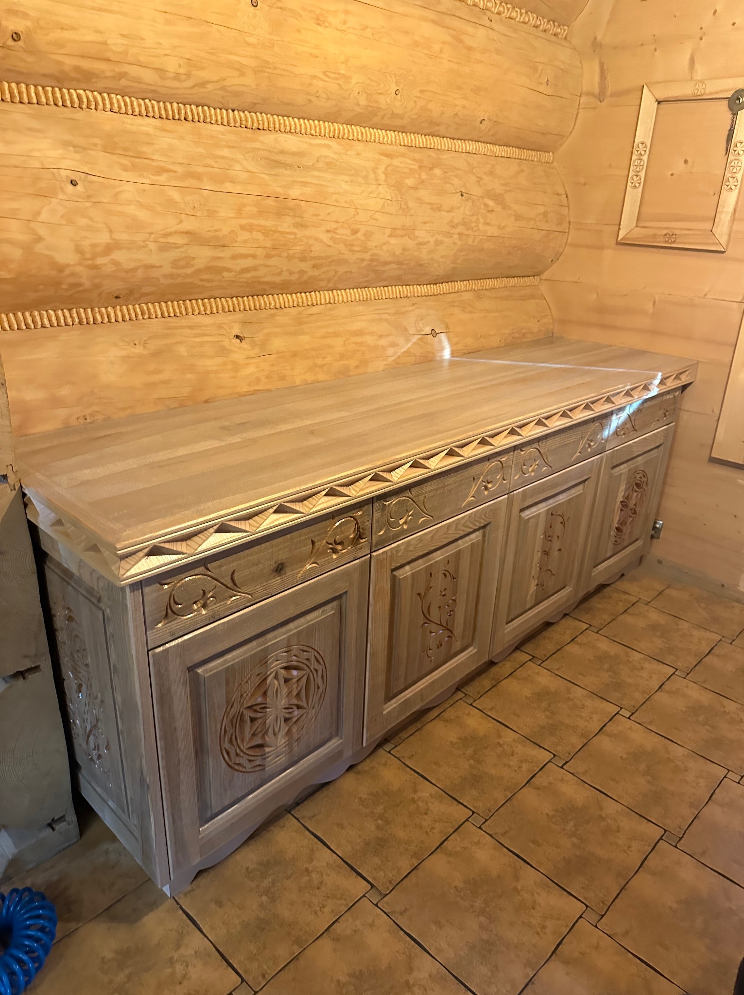 Drewniana komoda z rzeźbieniami w rustykalnym wnętrzu z drewnianych bali. Dekoracyjny element wykończenia i stylizowane płytki podłogowe.