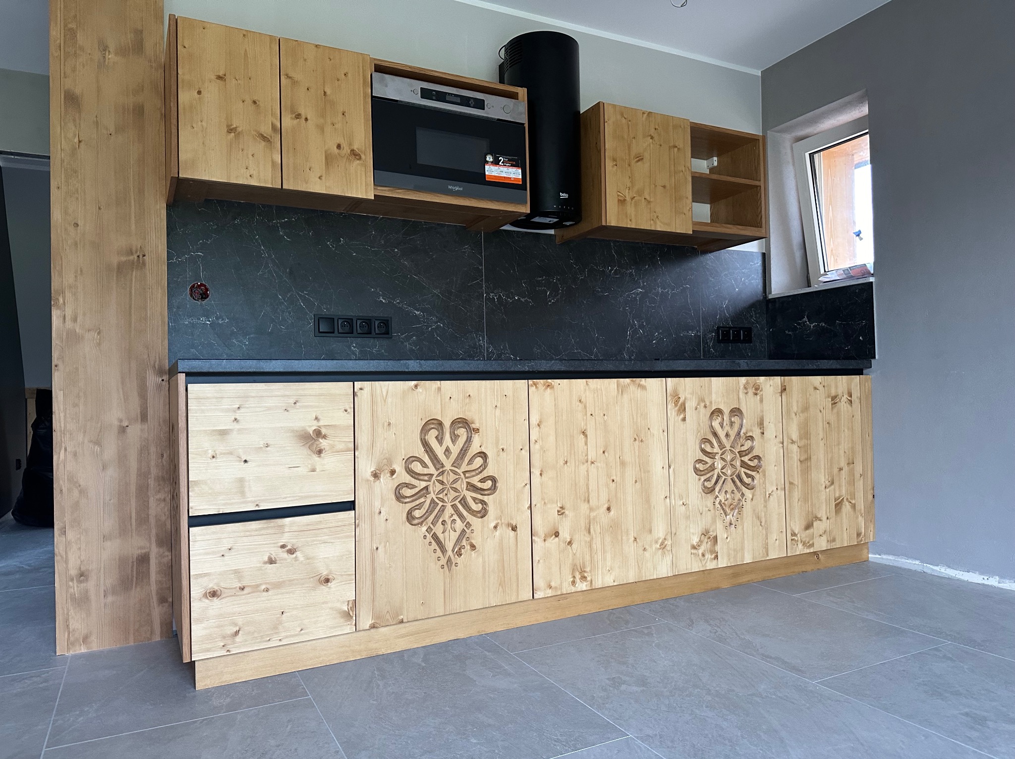 Drewniana kuchnia z frezowanymi wzorami, czarnym blatem i sprzętem AGD. Ujęcie z perspektywy podłogi, ukazujące detale wykończenia i nowoczesny design.