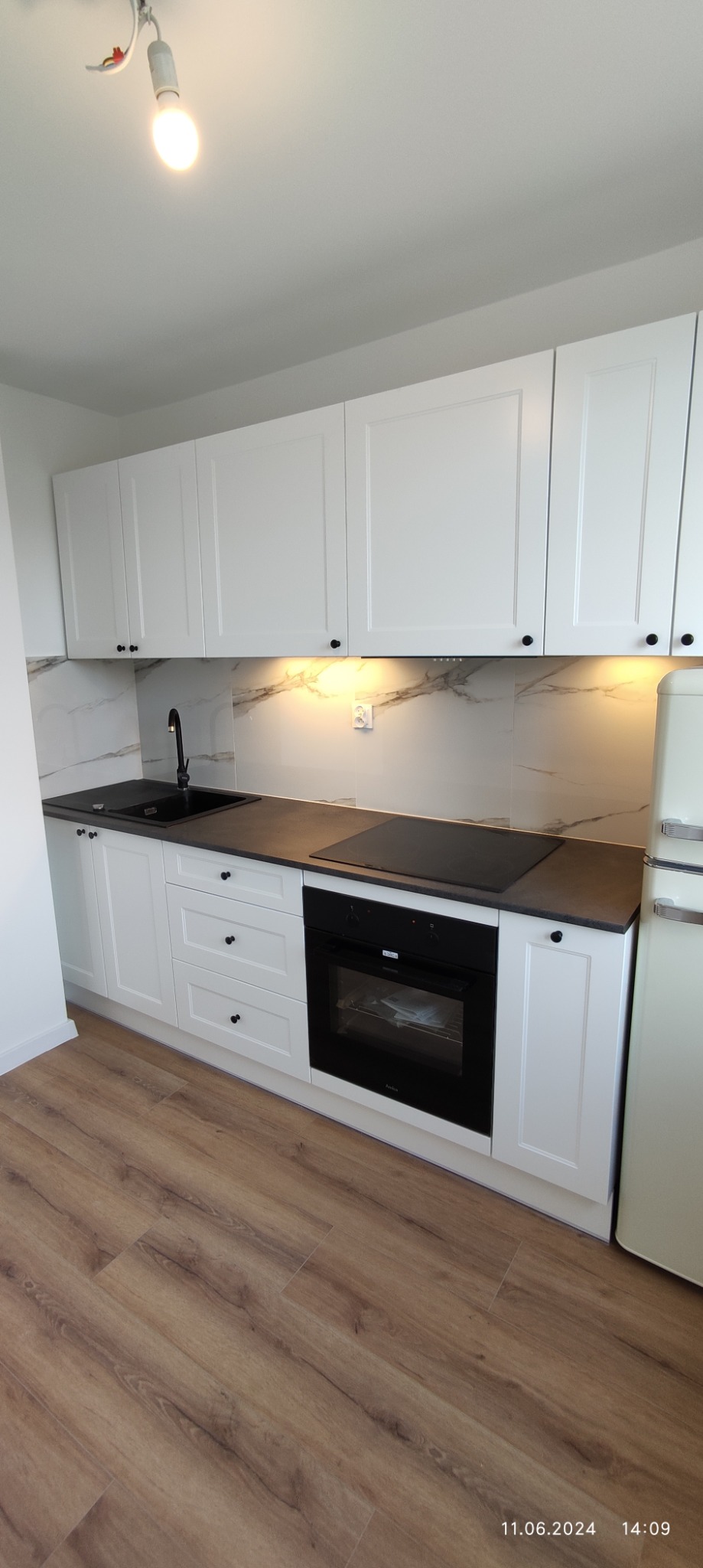 Nowoczesna, jasna kuchnia z białymi szafkami, czarnym blatem i marmurowym backsplash, z widoczną płytą indukcyjną i piekarnikiem.