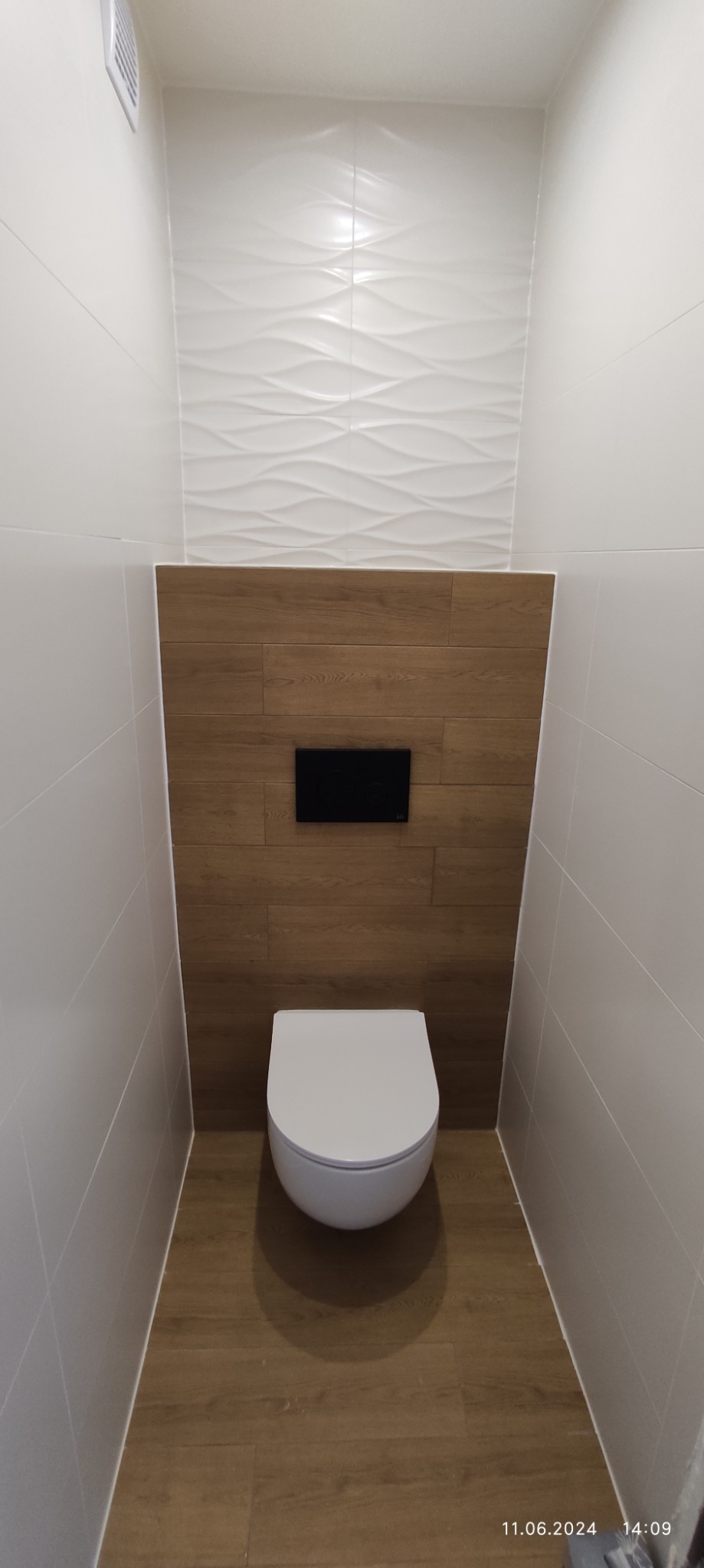 Nowoczesna toaleta podwieszana w wykończonej łazience z białymi i drewnopodobnymi płytkami, czarny panel sterujący, minimalistyczny design.