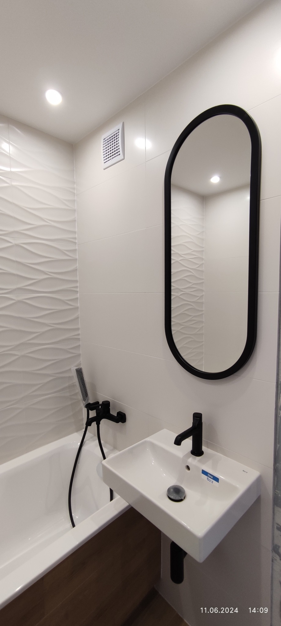 Nowoczesna łazienka z białą umywalką nablatową, czarną armaturą i owalnym lustrem w czarnej ramie, z dekoracyjnymi płytkami na ścianie i widoczną wanną.