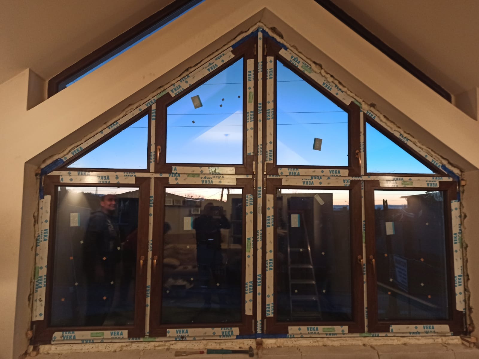 Montaż okien o nietypowym trójkątnym kształcie w nowym domu, widoczne zabezpieczenia folią i częściowo wykończone ściany, w tle drabina i sylwetki osób.