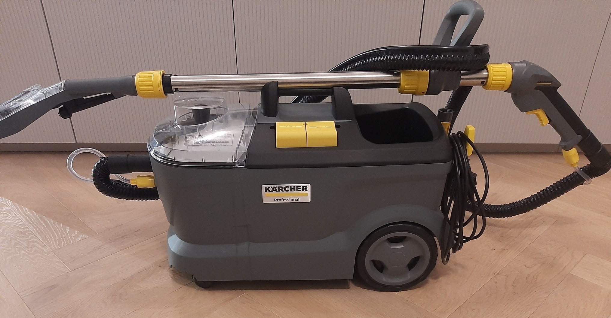 Profesjonalny odkurzacz piorący Kärcher z przezroczystym zbiornikiem na wodę, szarą obudową z żółtymi akcentami i licznymi wężami oraz dyszą, stojący na parkiecie.