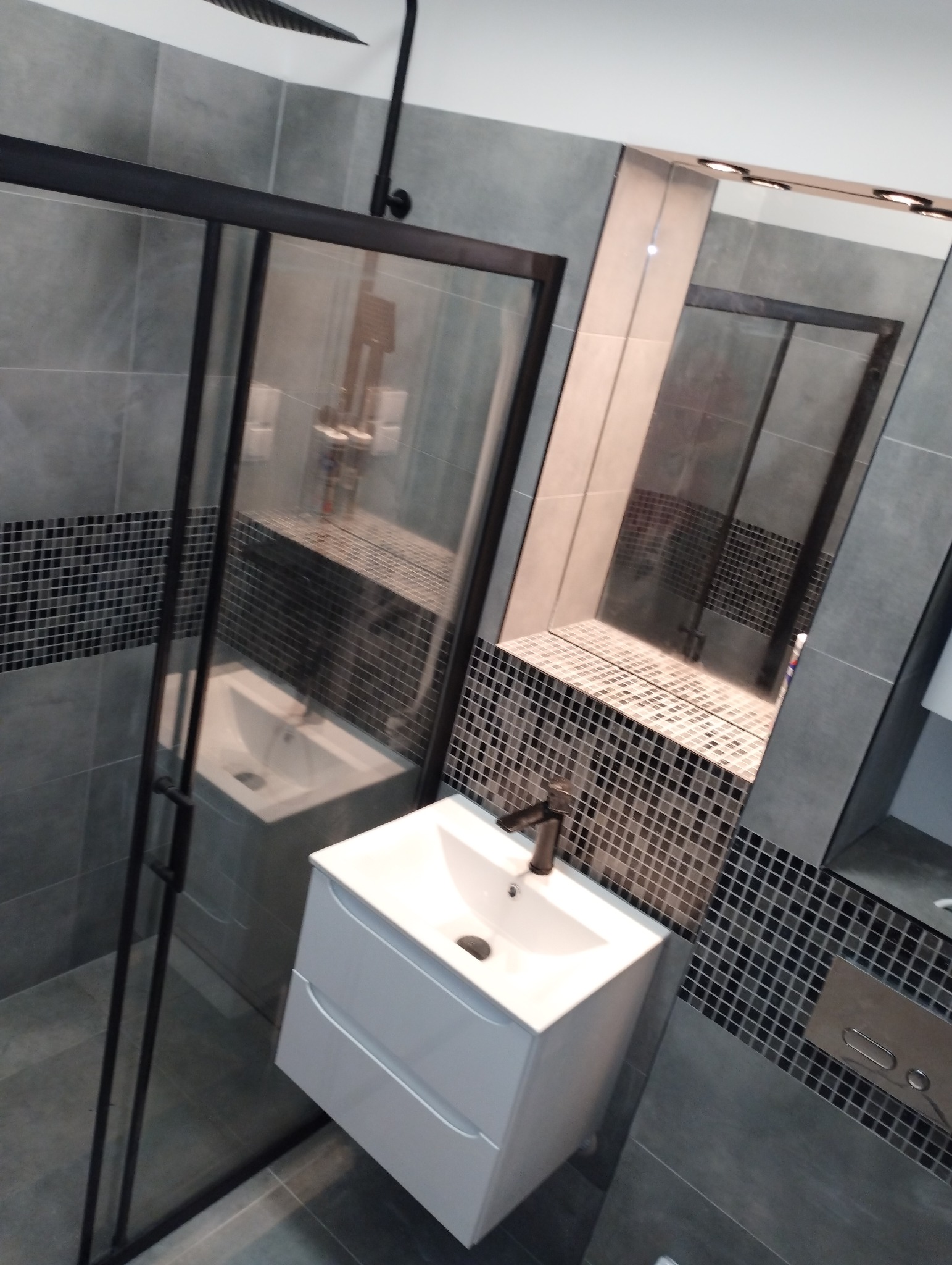 Nowoczesna łazienka z kabiną prysznicową typu walk-in z czarną ramą, białą szafką umywalkową z dwoma szufladami i lustrem z oświetleniem punktowym, wykończona szarą płytką imitującą beton i mozaiką...