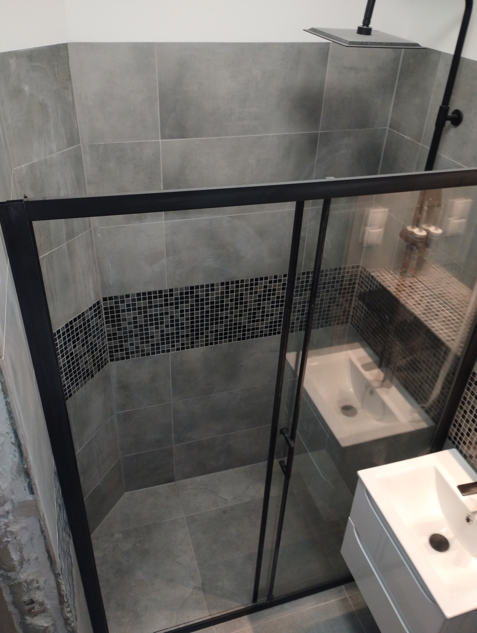 Nowoczesna kabina prysznicowa z czarnymi profilami i deszczownicą, szare płytki wielkoformatowe z pasem mozaiki, biała umywalka z szafką, wykończona łazienka.