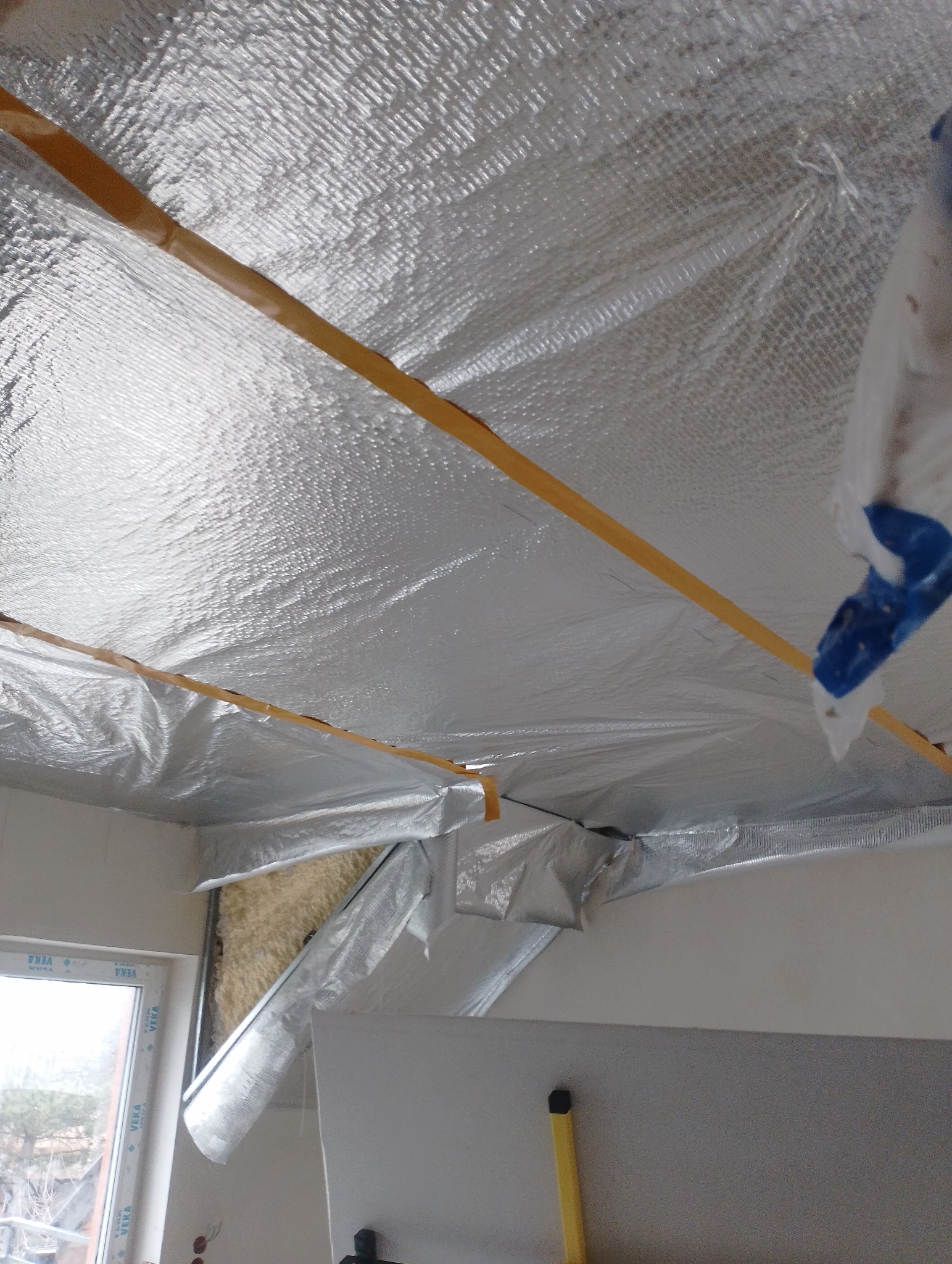 Ocieplenie poddasza folią aluminiową, widoczne fragmenty wełny mineralnej i okno PCV.