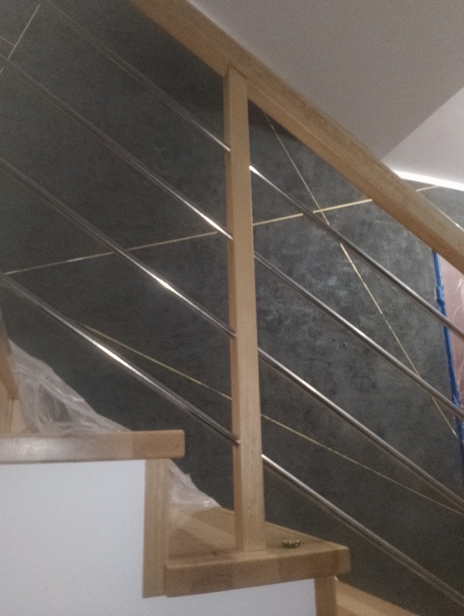 Fragment drewnianych schodów z metalowymi barierkami na tle ściany z szarego kamienia z delikatnym złotym wzorem.