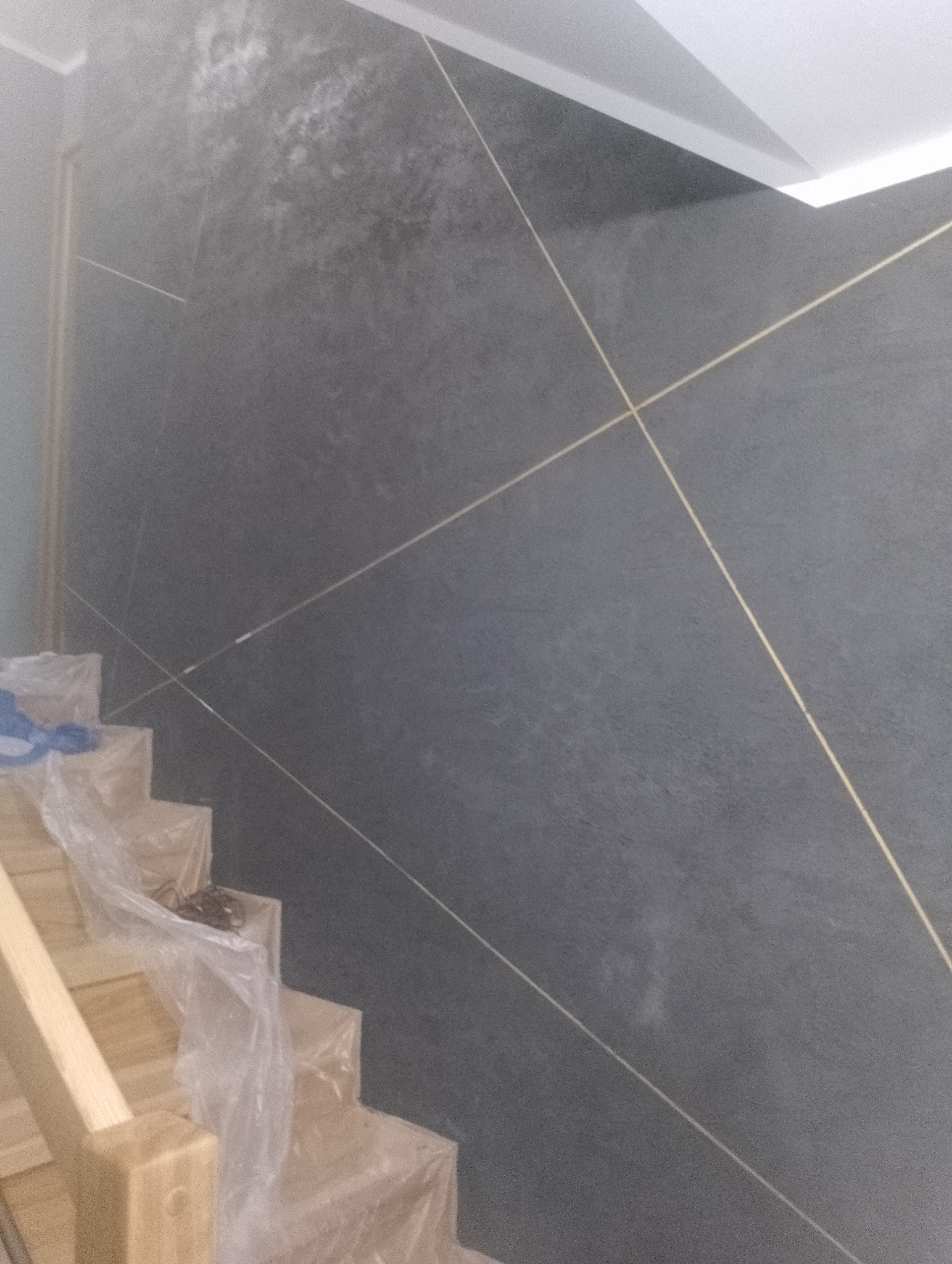 Ściana w odcieniach szarości z dekoracyjnymi, złotymi liniami, częściowo zasłonięta zabezpieczonymi folią schodami.