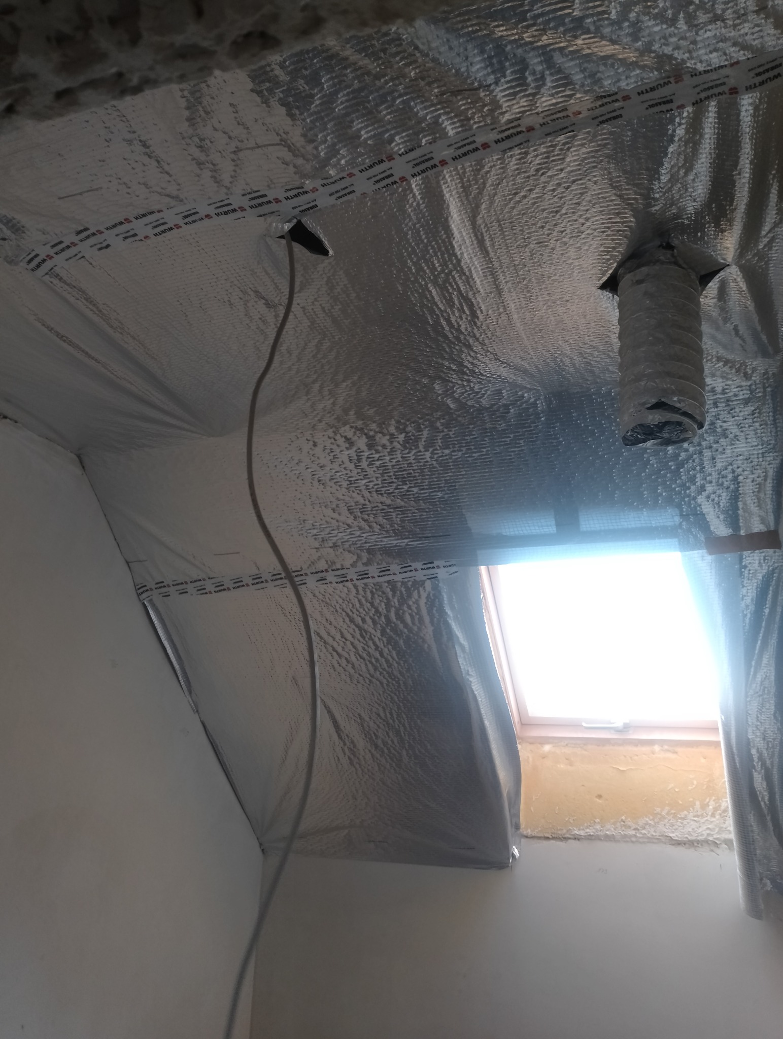 Montaż izolacji termicznej z folią aluminiową na poddaszu, widoczna rura wentylacyjna i kabel elektryczny, okno dachowe w tle.