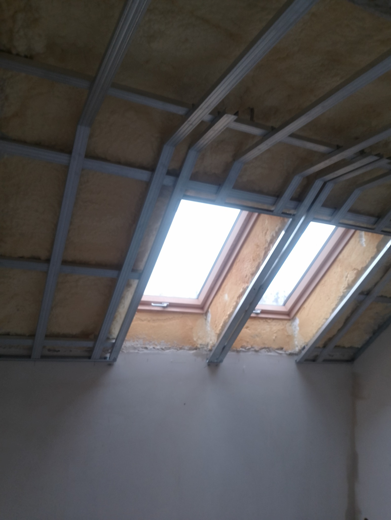 Skośny sufit poddasza z widoczną izolacją z wełny mineralnej między krokwiami i metalowym stelażem pod płyty gipsowo-kartonowe, dwa okna dachowe w drewnianych ramach wpuszczone w konstrukcję.