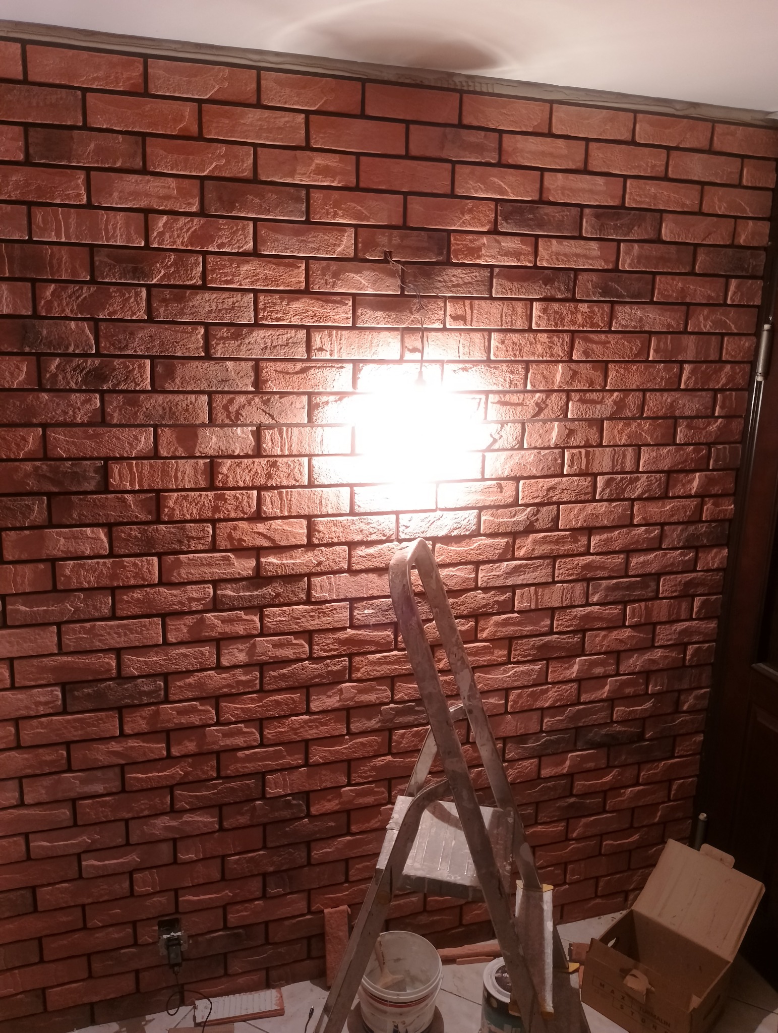 Wykończona ściana z dekoracyjnej cegły w odcieniach czerwieni i brązu, oświetlona centralnie, z widoczną drabiną i akcesoriami malarskimi na podłodze, sugerująca prace wykończeniowe.