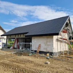 Budowa domu w trakcie realizacji: ściany z bloczków, czarny dach z dachówki, rusztowania. Widok na plac budowy w słoneczny dzień.