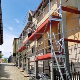 Elewacja domu w Zagórzanach w trakcie prac. Widoczne rusztowanie z pracownikiem. Dom ocieplany, z brązowym dachem. Ulica wyłożona kostką.