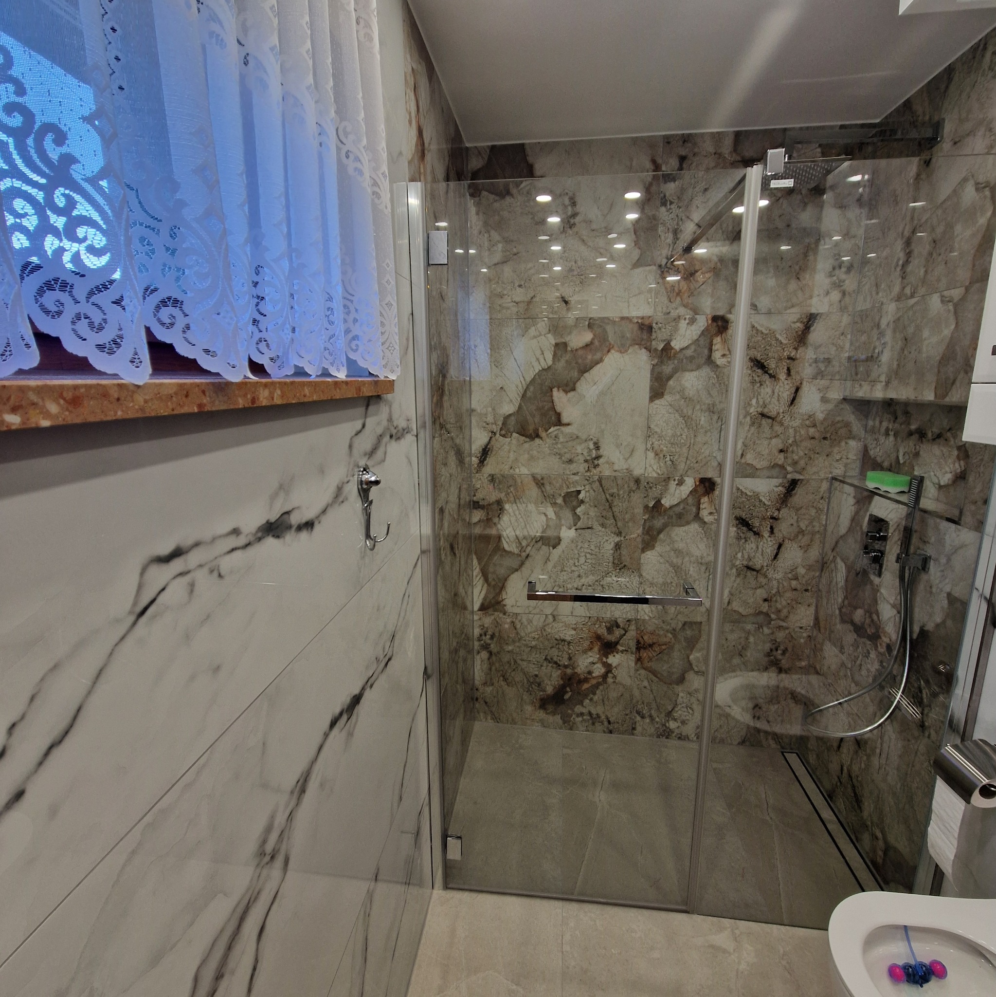 Wykończona łazienka z kabiną prysznicową z marmurowymi płytkami i ozdobną zasłoną w oknie. Widoczny fragment toalety. Nowoczesny design.