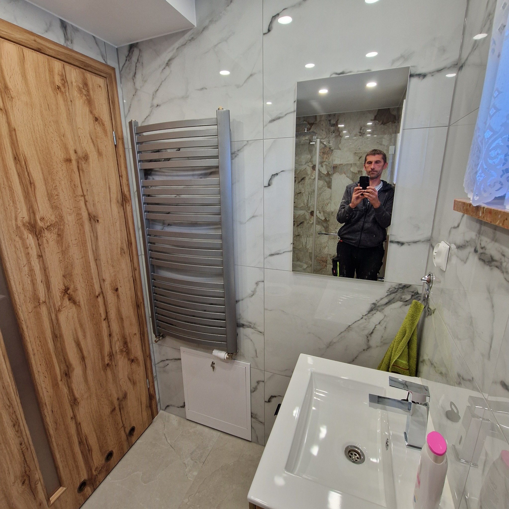 Wykończona łazienka w bieli i szarości z marmurowym wzorem. Widoczna umywalka, grzejnik drabinkowy i drewniane drzwi. W lustrze odbicie osoby robiącej zdjęcie.