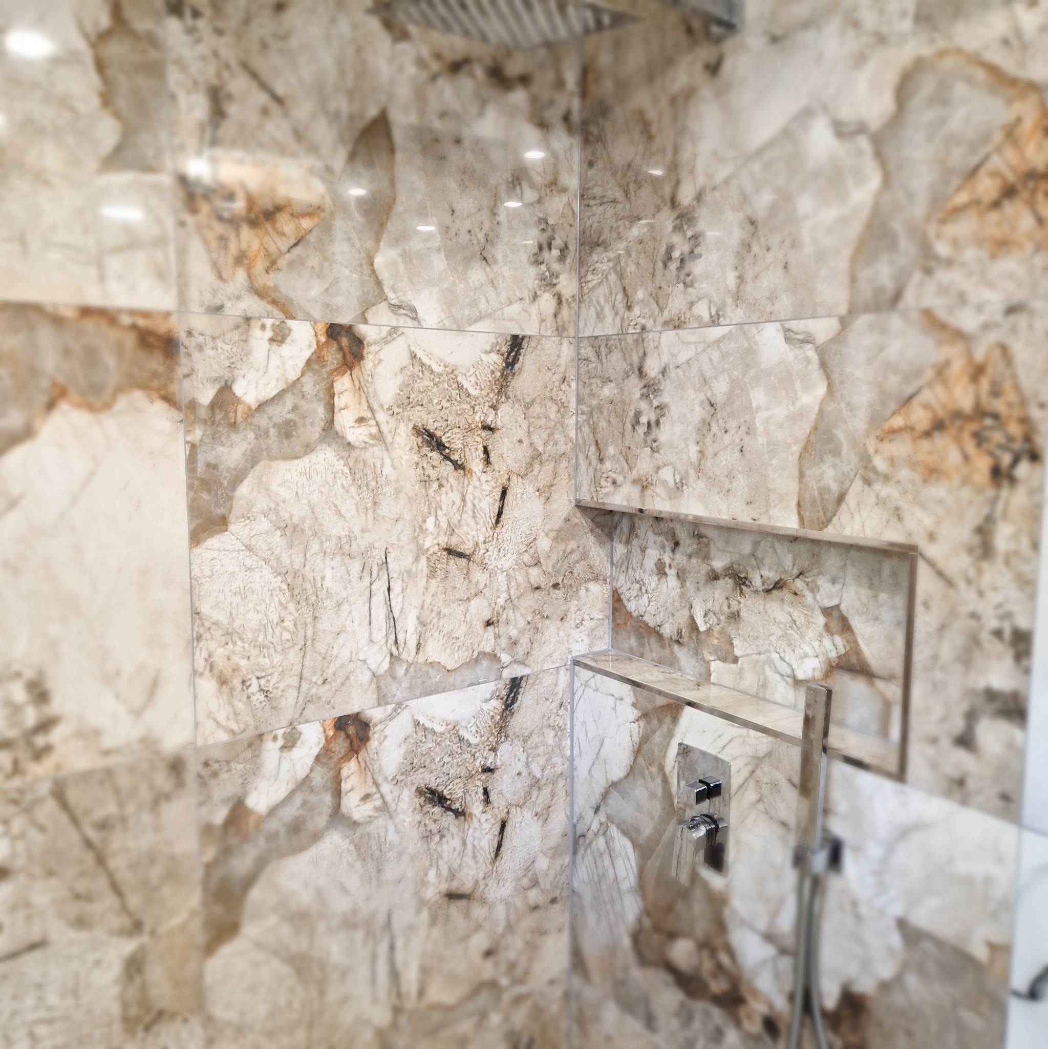 Wykończona kabina prysznicowa z marmurowymi płytkami w odcieniach beżu i brązu. Wbudowane półki i nowoczesna armatura. Widok z dołu na górę.