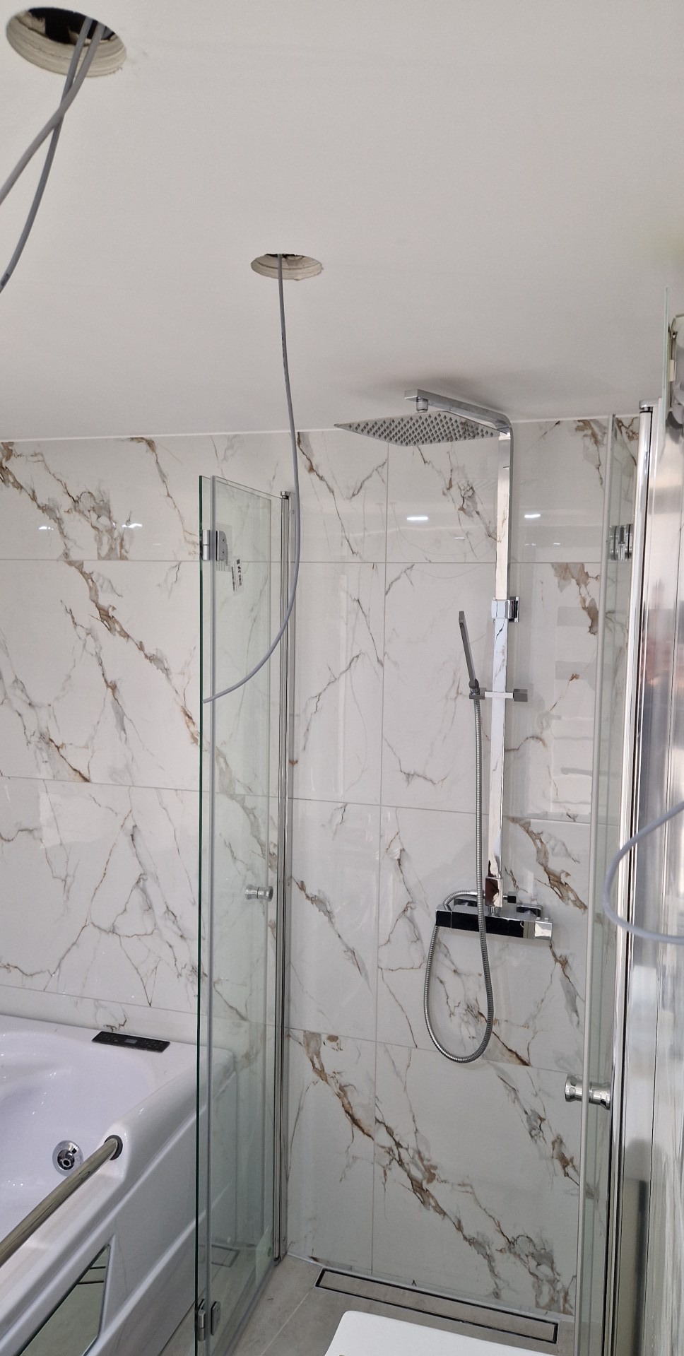 Nowoczesna kabina prysznicowa z deszczownicą i marmurowymi płytkami. Widoczne niedokończone podłączenia oświetlenia sufitowego.