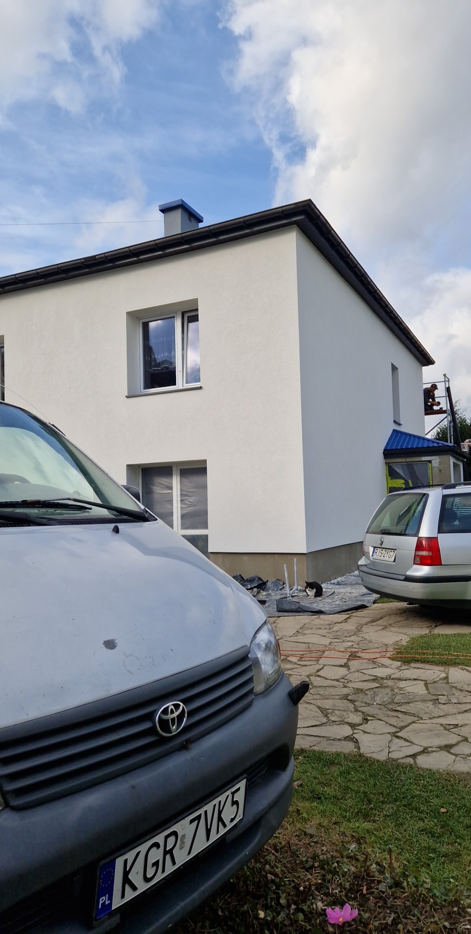 Biała elewacja dwupiętrowego domu po tynkowaniu, z widocznymi dwoma samochodami i kotem siedzącym na folii ochronnej. W tle pracownik na rusztowaniu.