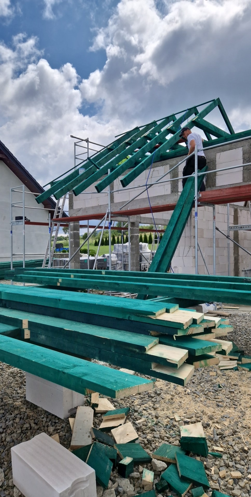 Wznoszenie konstrukcji dachu na budynku w trakcie budowy. Widoczne zielone belki, rusztowania i pracownik na wysokości. W tle pochmurne niebo.