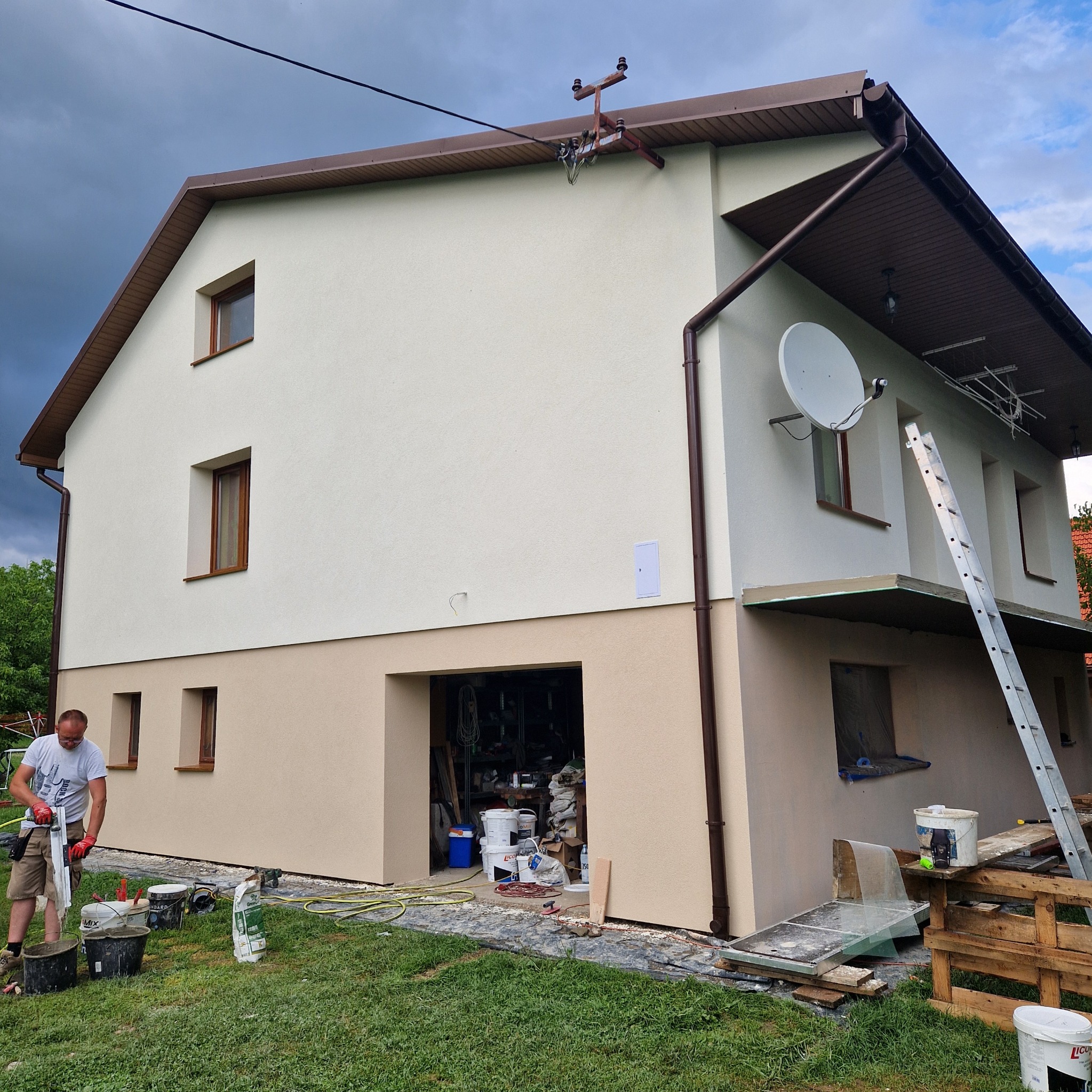 Dwukondygnacyjny dom po tynkowaniu elewacji, z widocznym pracownikiem mieszającym zaprawę. Ściany w odcieniach beżu, brązowe okna i dach. Na elewacji antena satelitarna.