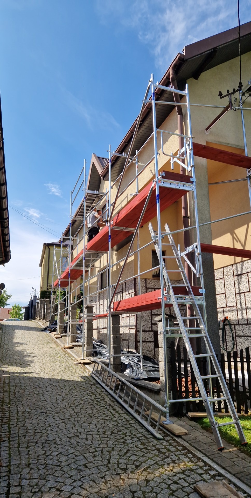 Elewacja domu w Zagórzanach w trakcie prac. Widoczne rusztowanie z pracownikiem. Dom ocieplany, z brązowym dachem. Ulica wyłożona kostką.