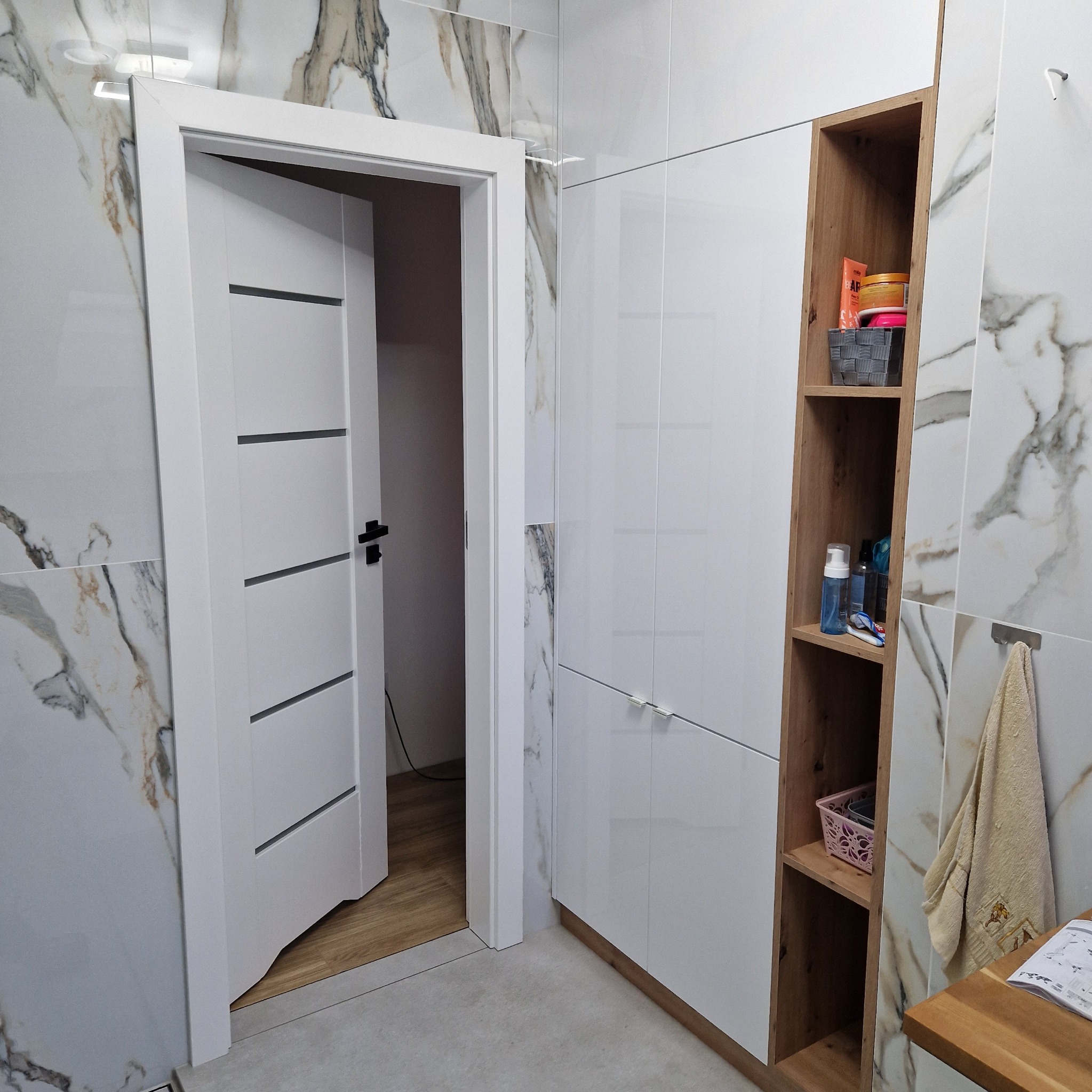 Nowoczesna łazienka po remoncie: białe płytki imitujące marmur, białe szafki z drewnianymi akcentami, otwarte półki i białe drzwi z czarną klamką.