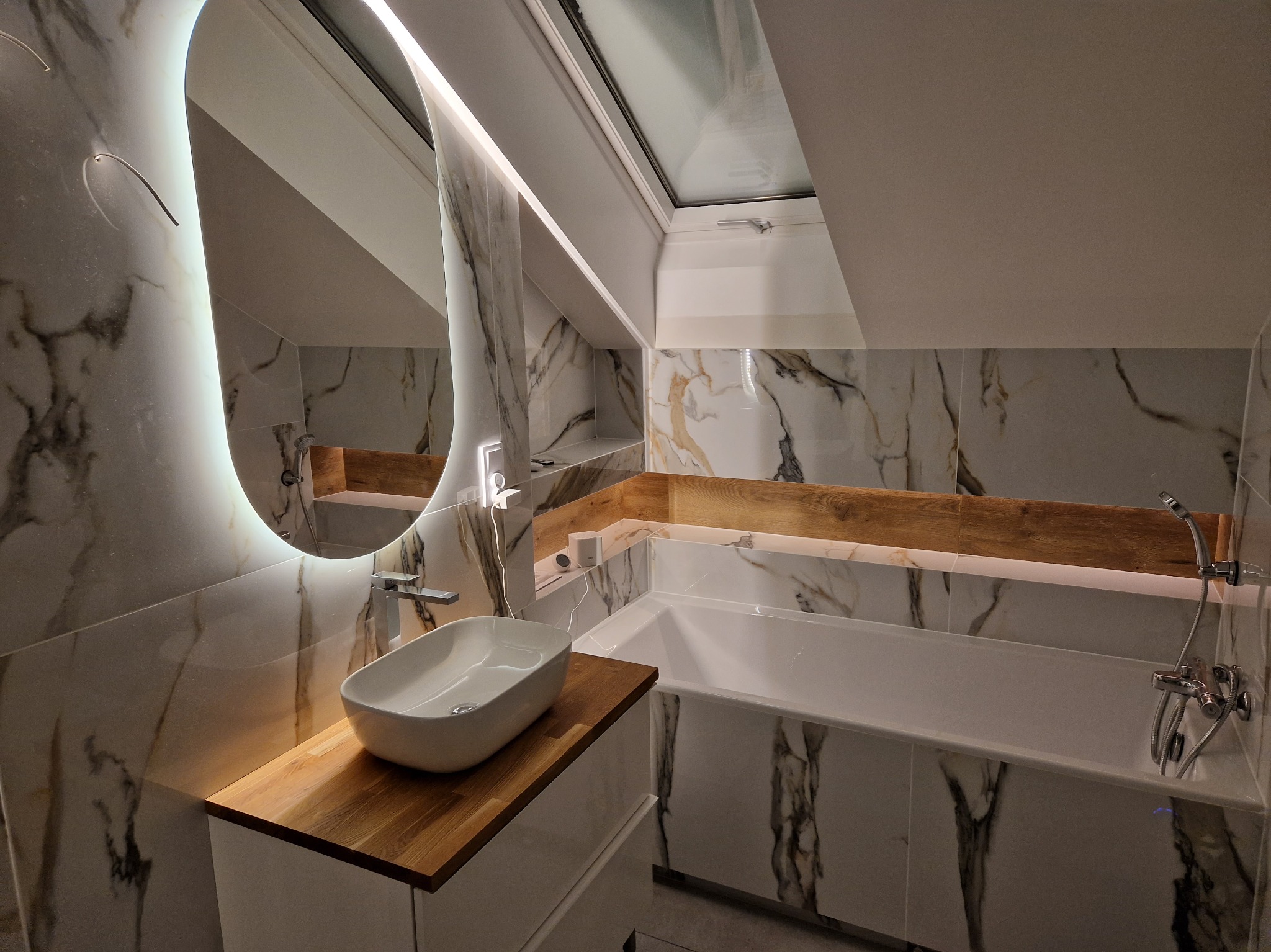 Nowoczesna łazienka na poddaszu z marmurowymi płytkami, owalnym lustrem z podświetleniem LED i drewnianymi akcentami. Umywalka nablatowa i wanna w zabudowie.