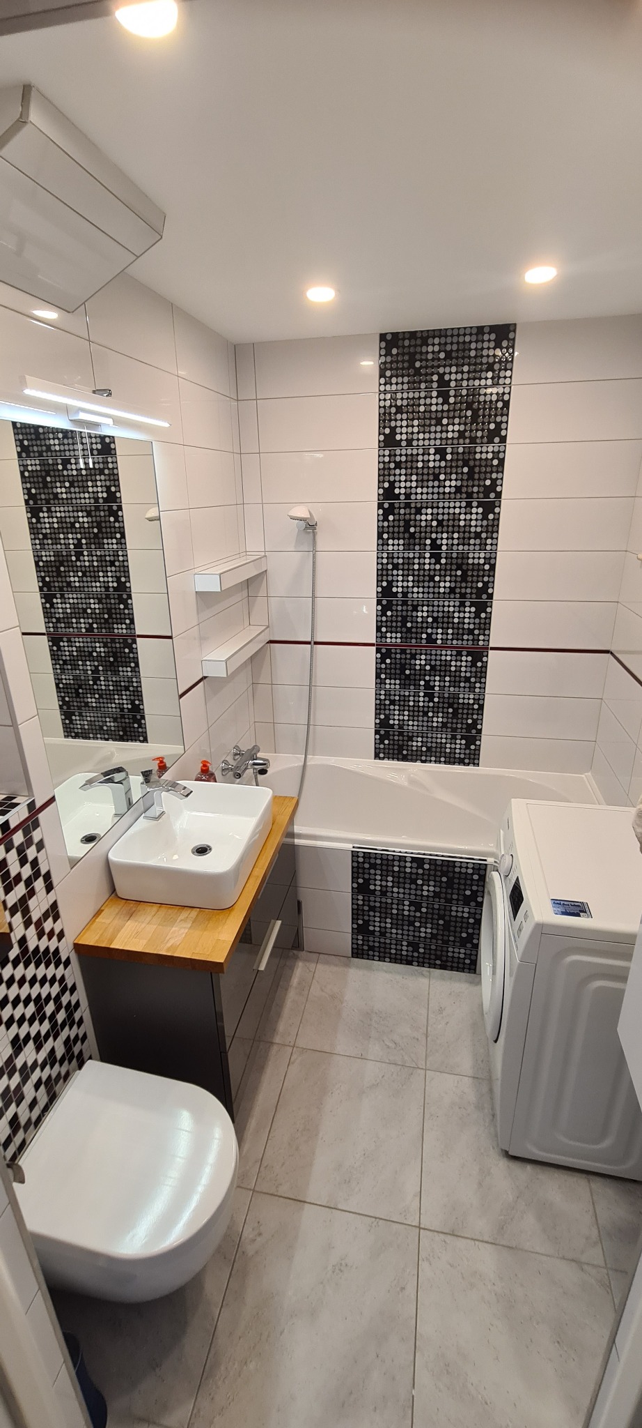 Nowoczesna łazienka z białymi i czarnymi płytkami mozaikowymi, drewnianym blatem umywalki i pralką. Minimalistyczny design z akcentami kolorystycznymi.