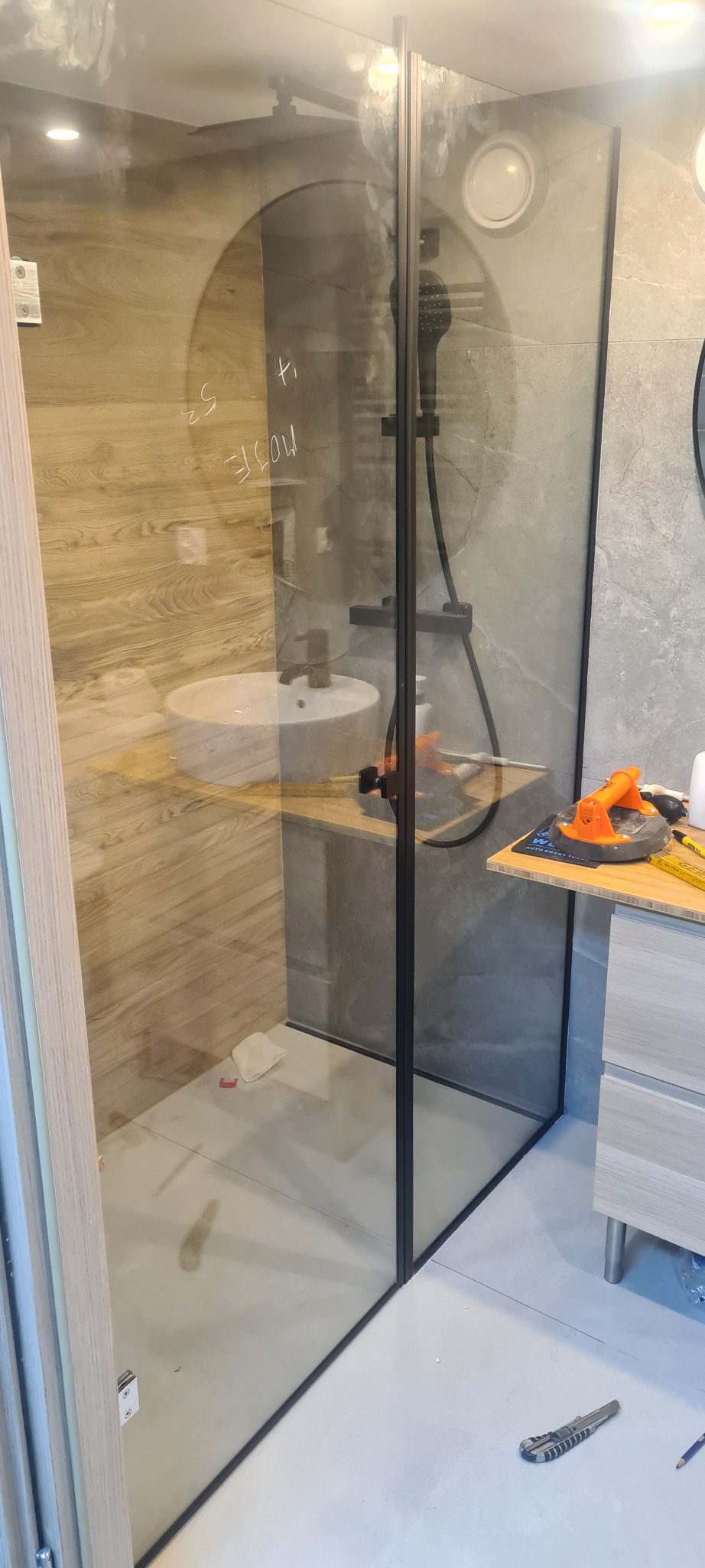 Nowoczesna łazienka z kabiną prysznicową typu walk-in, wykończona w odcieniach szarości i drewna. Widoczne narzędzia wskazują na trwające prace wykończeniowe.