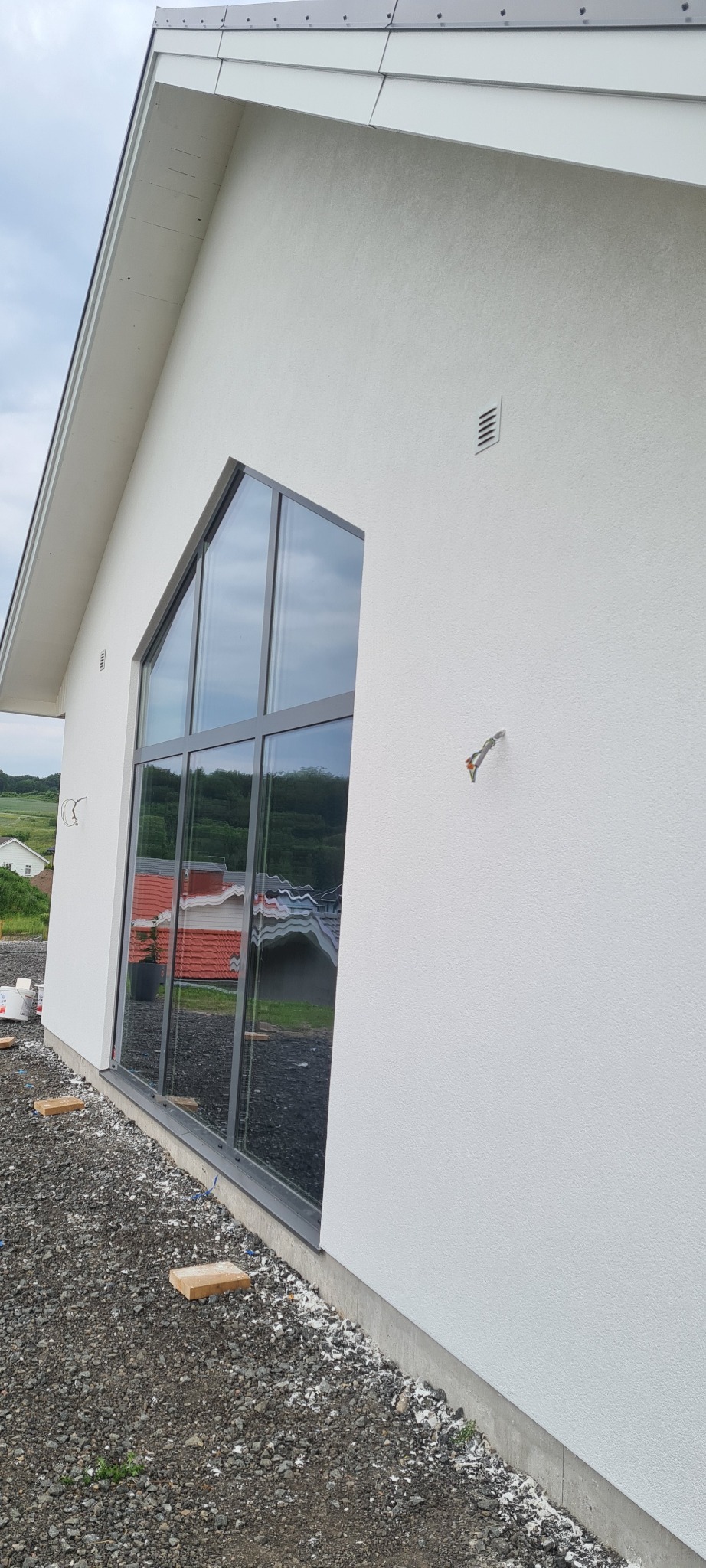 Biała elewacja domu z dużym oknem w kształcie trapezu. Widoczne detale wykończenia i struktura tynku. Na ziemi gruz i fragmenty materiałów budowlanych.