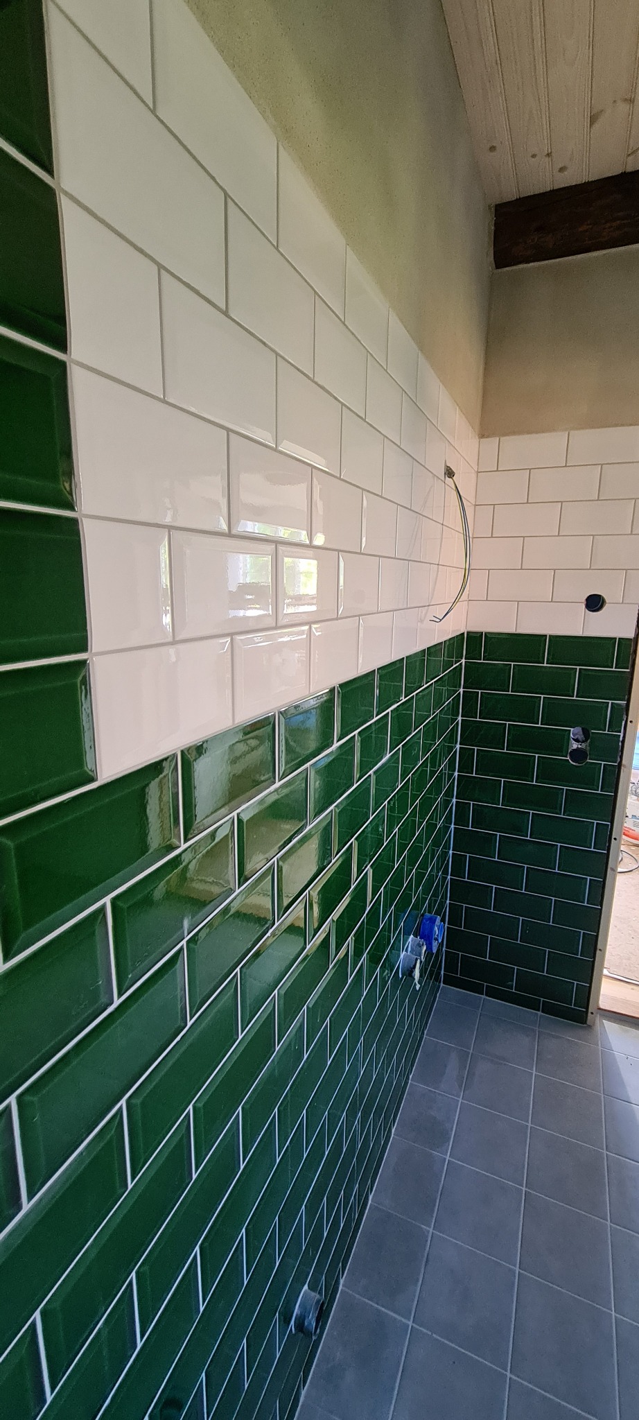 Ściana wyłożona błyszczącymi, zielonymi i białymi płytkami typu metro w łazience. Widoczne szare płytki podłogowe i fragment sufitu z drewnianą boazerią.