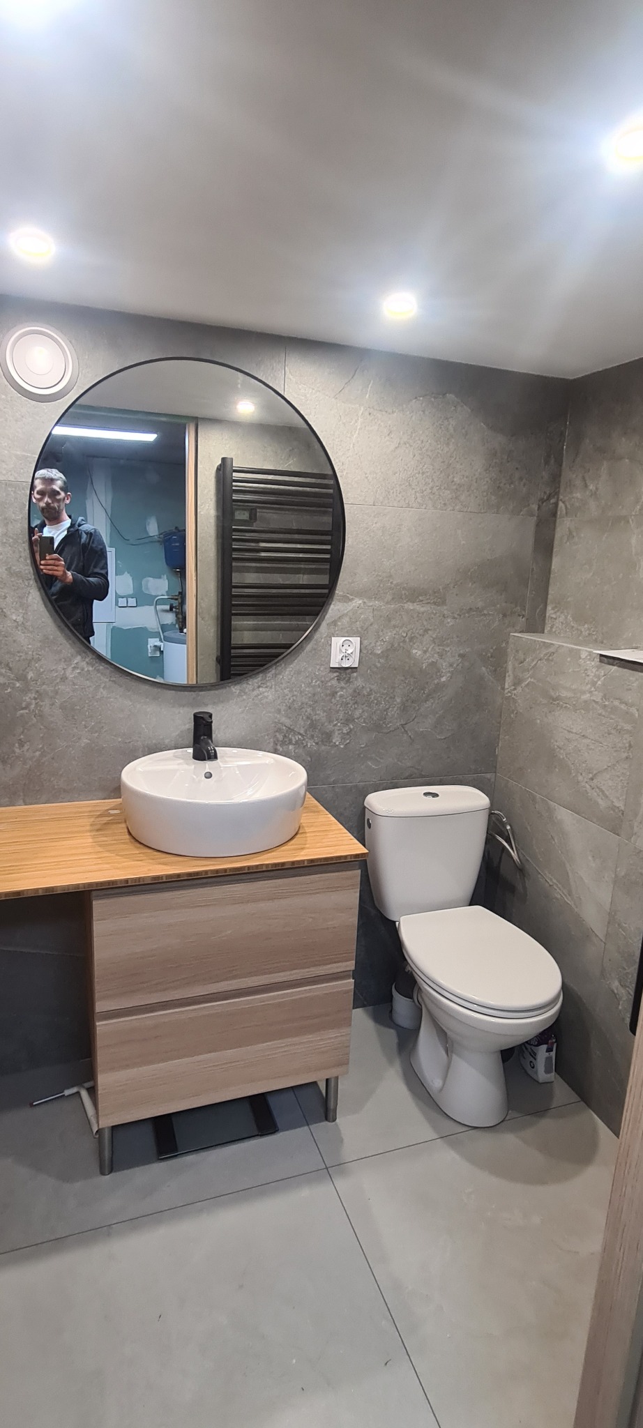 Nowoczesna łazienka z umywalką nablatową, okrągłym lustrem z odbiciem osoby, szarą płytką i wiszącą grzejnikiem. Minimalistyczny design.