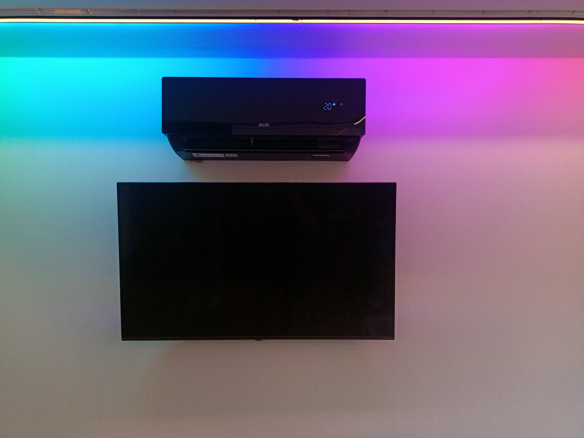 Czarny klimatyzator marki AUX zamontowany na ścianie nad czarnym telewizorem, oświetlony z góry paskiem LED zmieniającym kolory od turkusowego do różowego, ustawiony na 20 stopni Celsjusza.