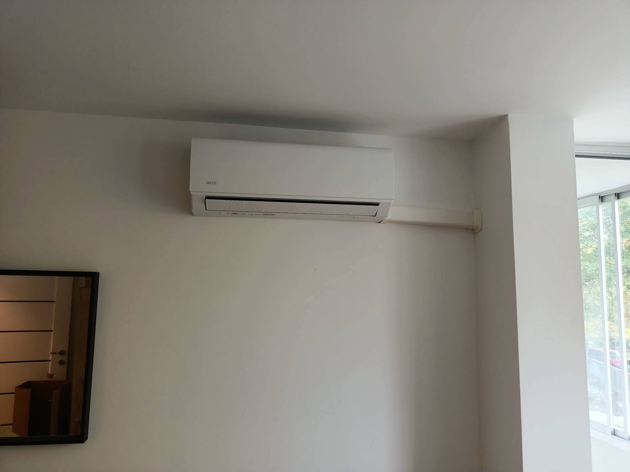 Biały klimatyzator marki AUX zamontowany na białej ścianie w pomieszczeniu z oknem i lustrem.