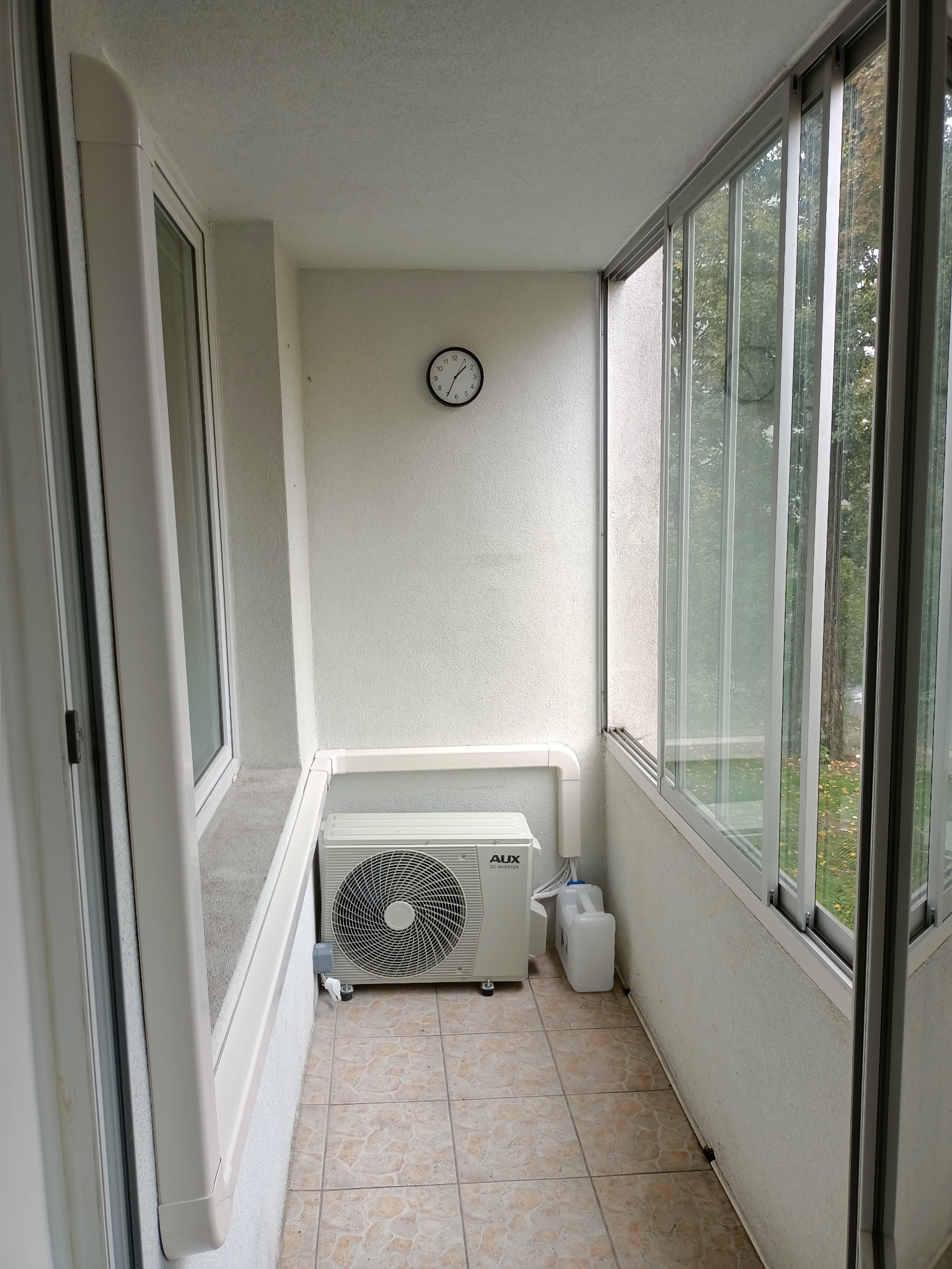 Biały klimatyzator AUX DC Inverter zamontowany na balkonie z przesuwnymi oknami, widoczny zegar ścienny i zbiornik na skropliny.