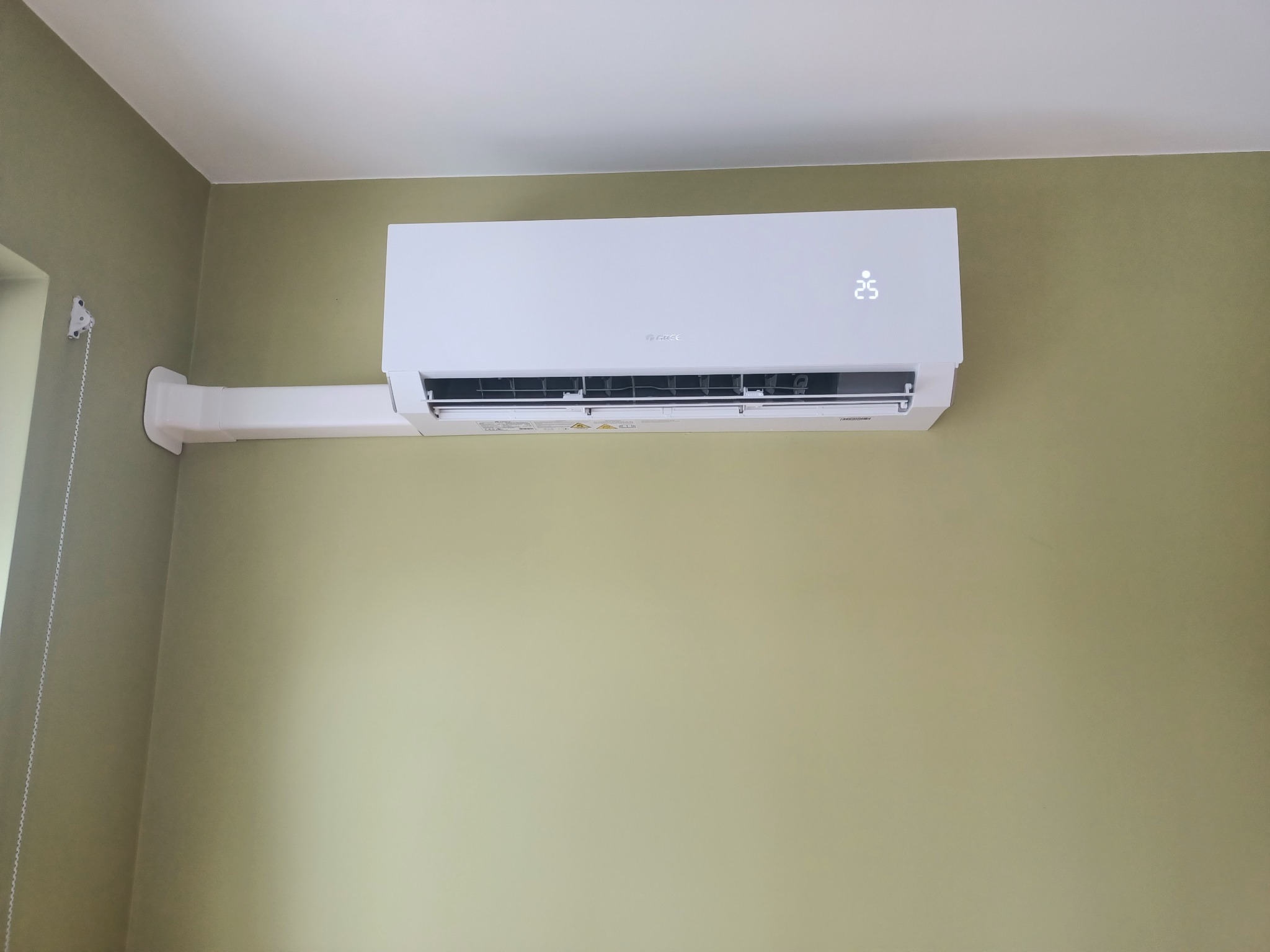 Biały klimatyzator marki Gree zamontowany na oliwkowozielonej ścianie, widoczna temperatura 25 stopni Celsjusza na wyświetlaczu.