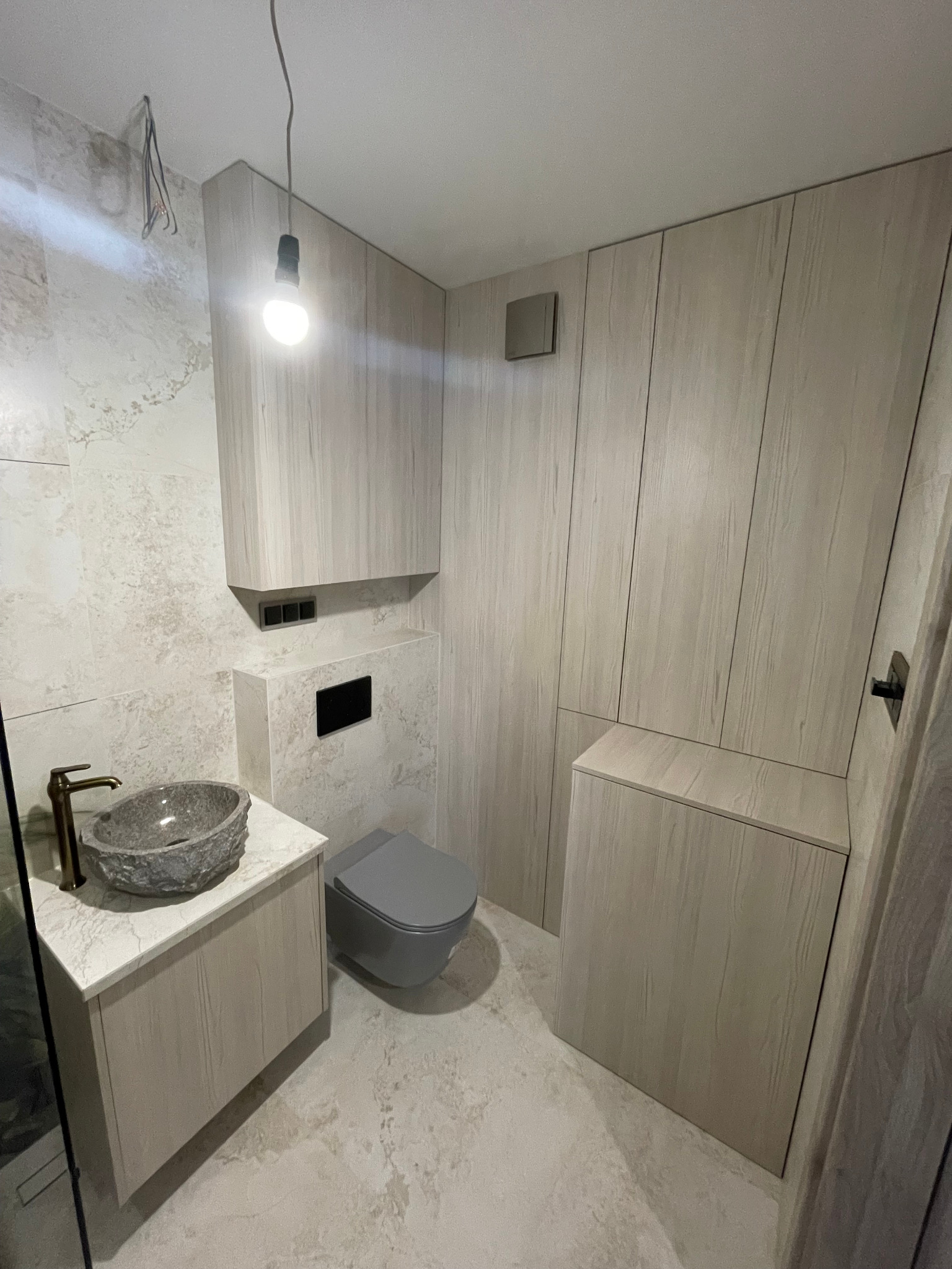 Nowoczesna łazienka z szafkami na wymiar w odcieniach beżu i szarości. Umywalka z kamienia, podwieszana toaleta, minimalistyczne oświetlenie. Całość tworzy spójny, elegancki design.