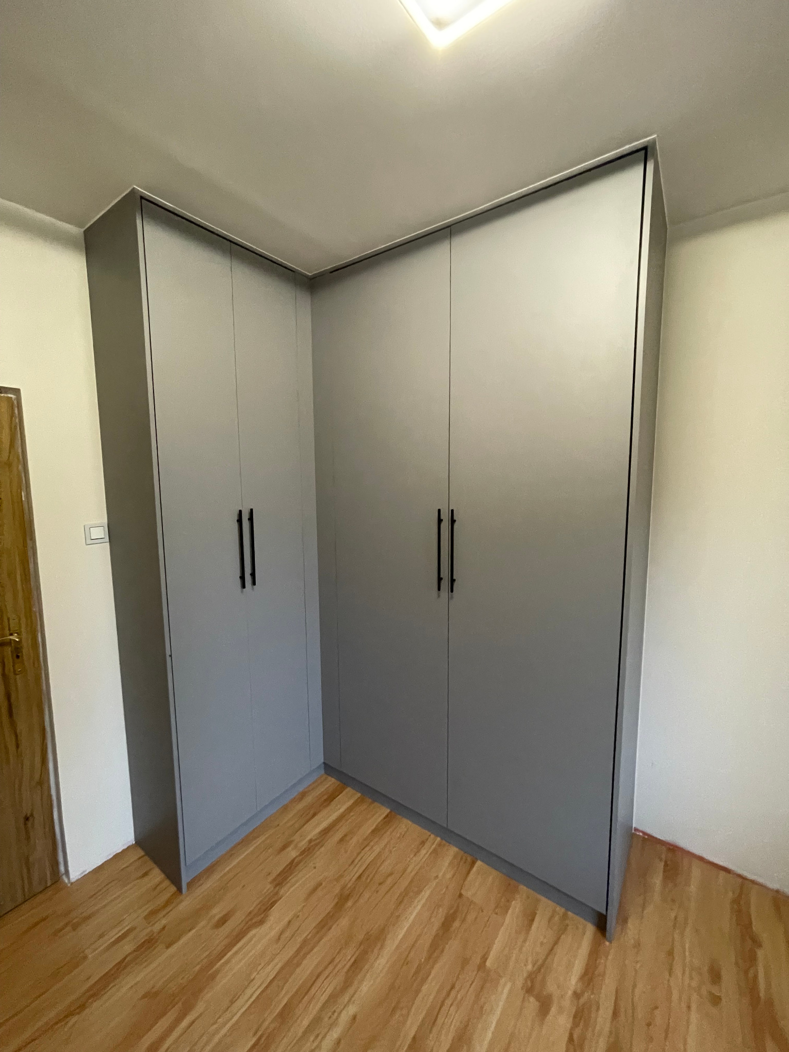 Szara szafa narożna z czarnymi, pionowymi uchwytami, dopasowana do skosu pomieszczenia, na drewnianej podłodze. Minimalistyczny design, proste linie.