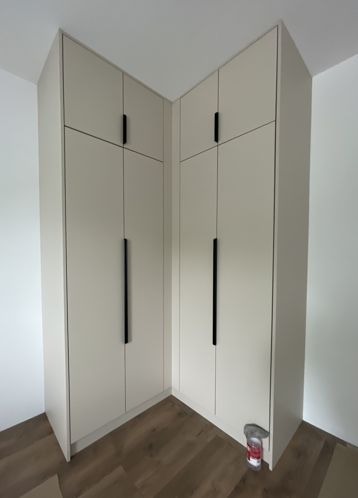 Jasna, narożna szafa z czarnymi uchwytami, dopasowana do wnętrza. Widoczna butelka z płynem do czyszczenia przy podstawie szafy. Podłoga drewniana.