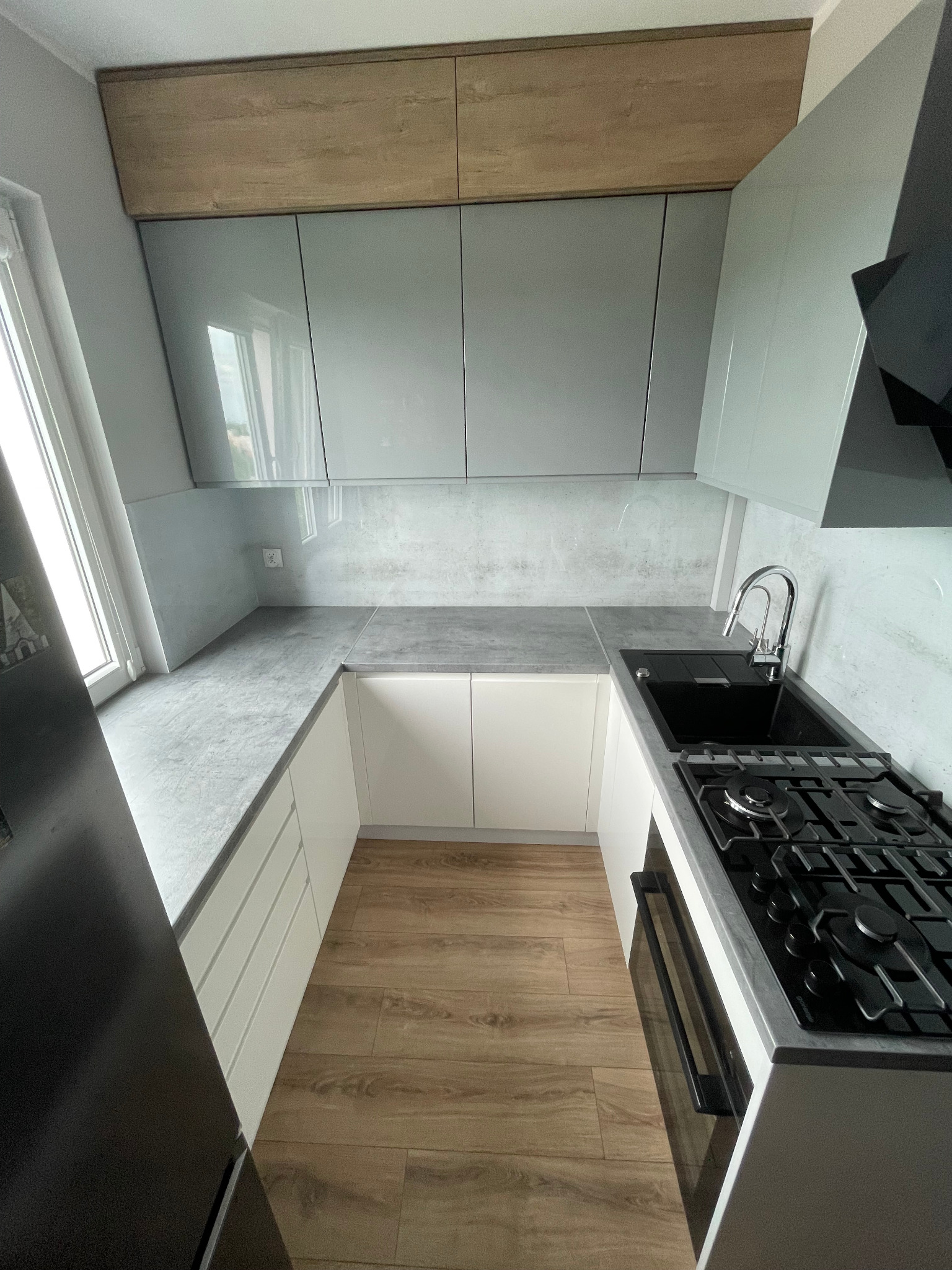 Nowoczesna kuchnia z szarymi szafkami górnymi i białymi dolnymi, drewnianą podłogą i czarną płytą grzewczą. Ujęcie z góry podkreśla układ i wykończenie.