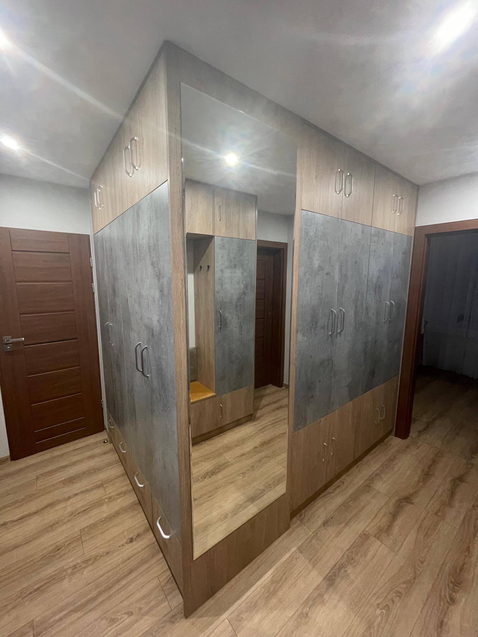 Szafa wnękowa z lustrem i szarymi frontami, drewnopodobne wykończenie, srebrne uchwyty, widok na wnętrze szafy w odbiciu lustra, drewniana podłoga.