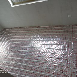 Michał Kulwicki Usługi hydrauliczne - Białe rury ogrzewania podłogowego ułożone na folii z czerwoną siatką na przygotowanym podłożu, widok z góry, surowy stan wykończenia wnętrza.